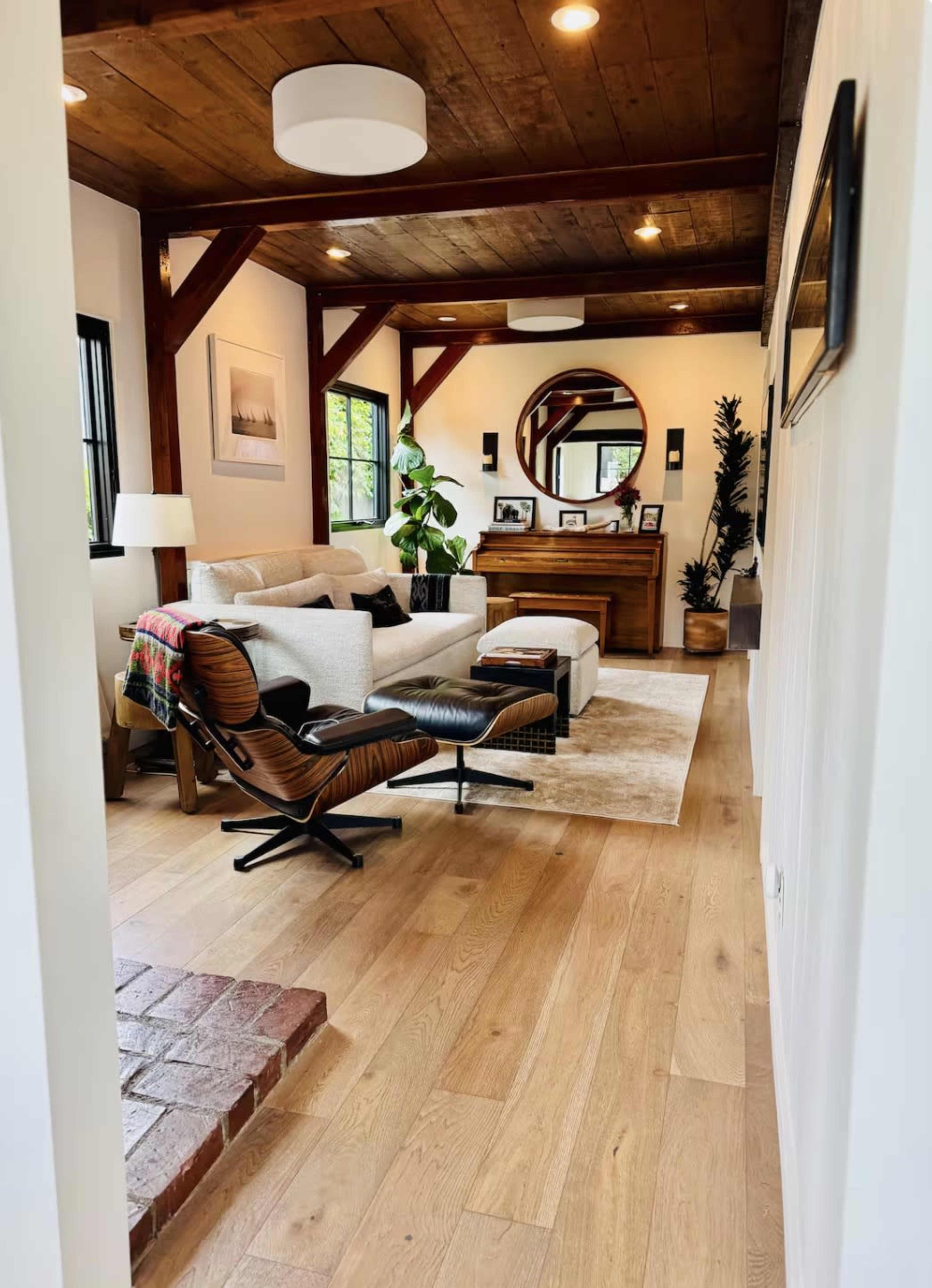A well-lit living room features a white sofa, a patterned throw, a wooden wall unit, and a round mirror, with wood beams on the ceiling and a view of a cozy entryway.
