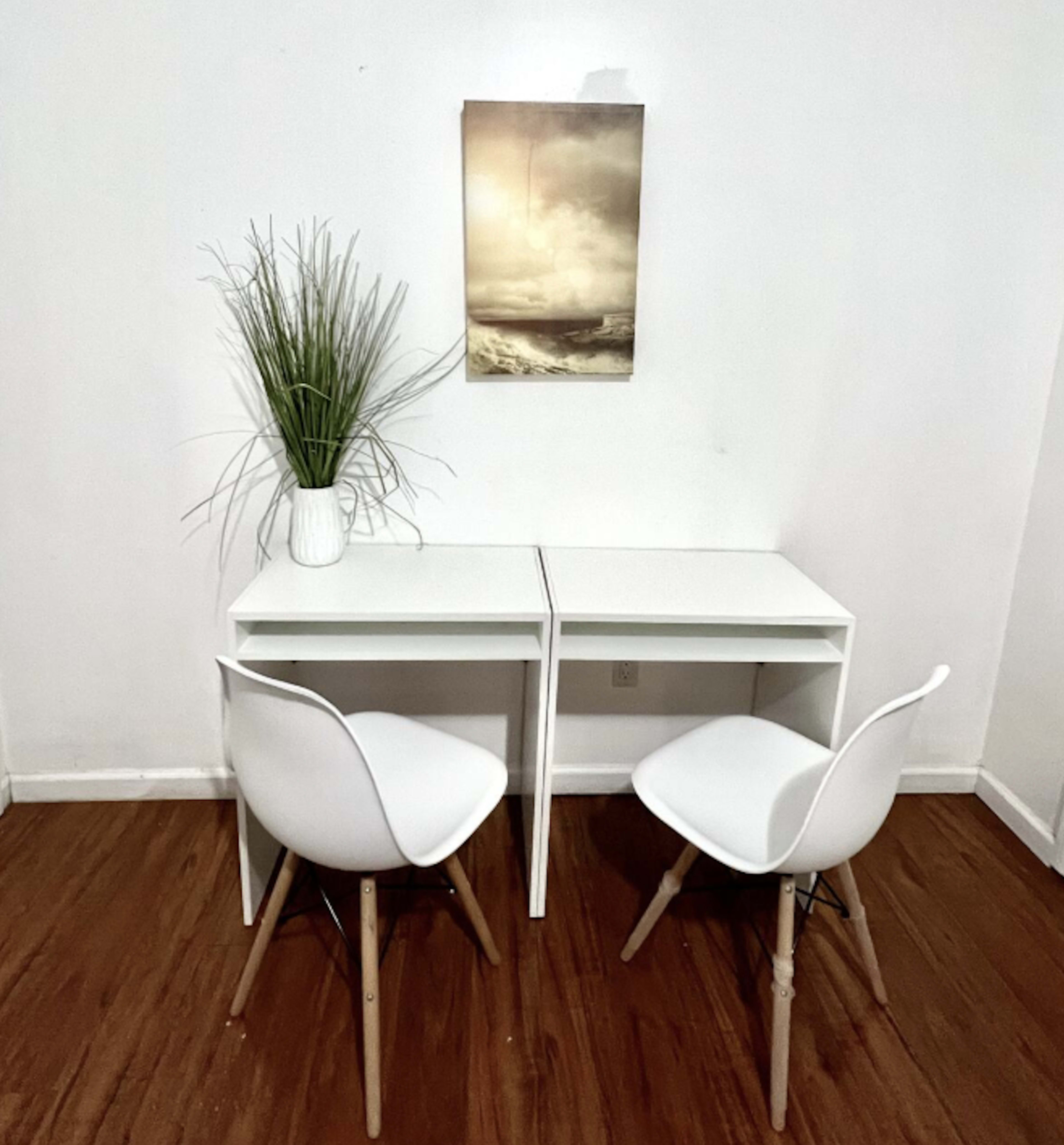 A simple white desk with two chairs sits against a plain wall, with a potted plant and a framed picture of a seascape above it.