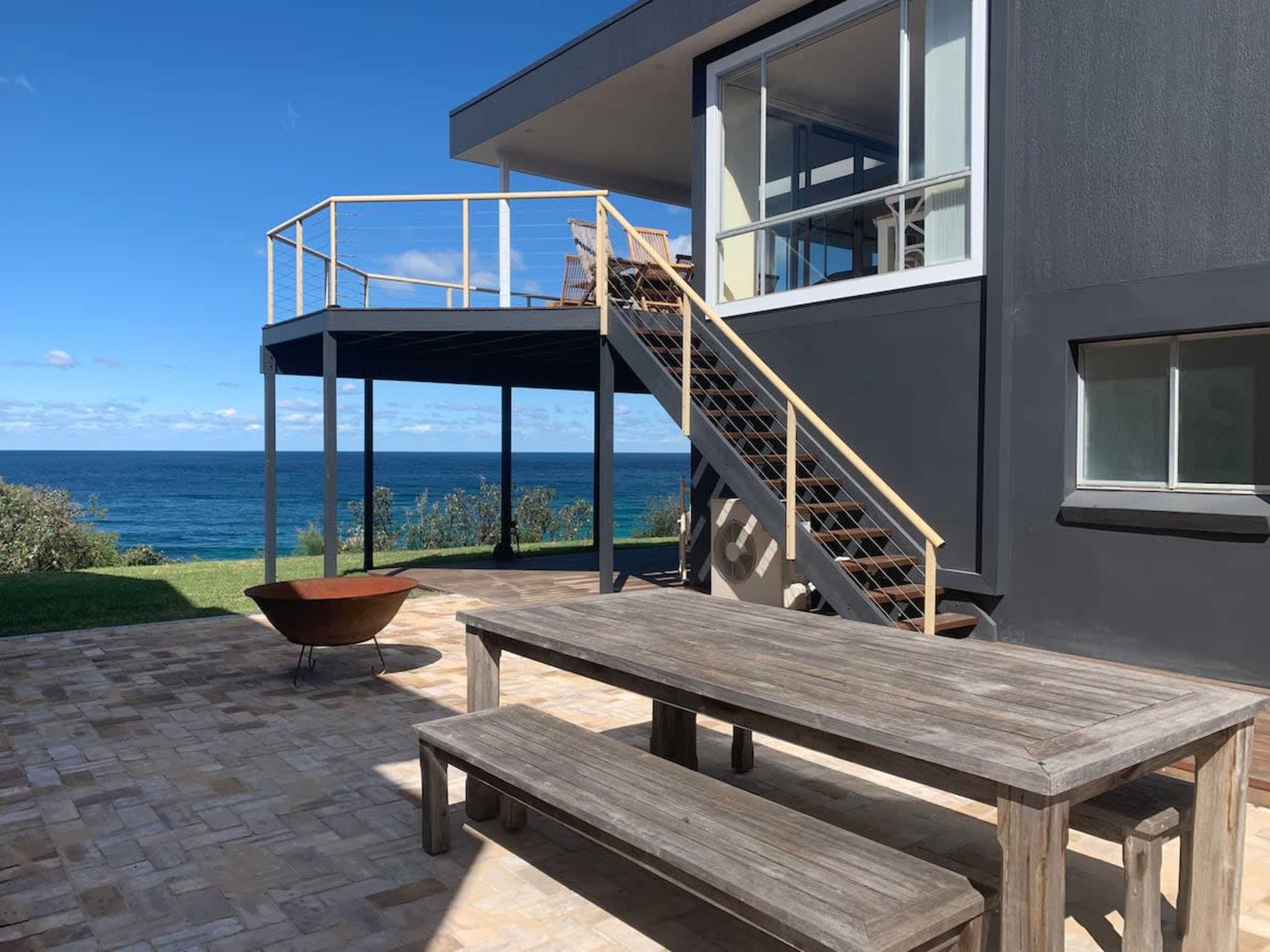 A wooden table and benches are set on a stone patio next to a modern house with a staircase leading to an upper deck and views of the ocean.