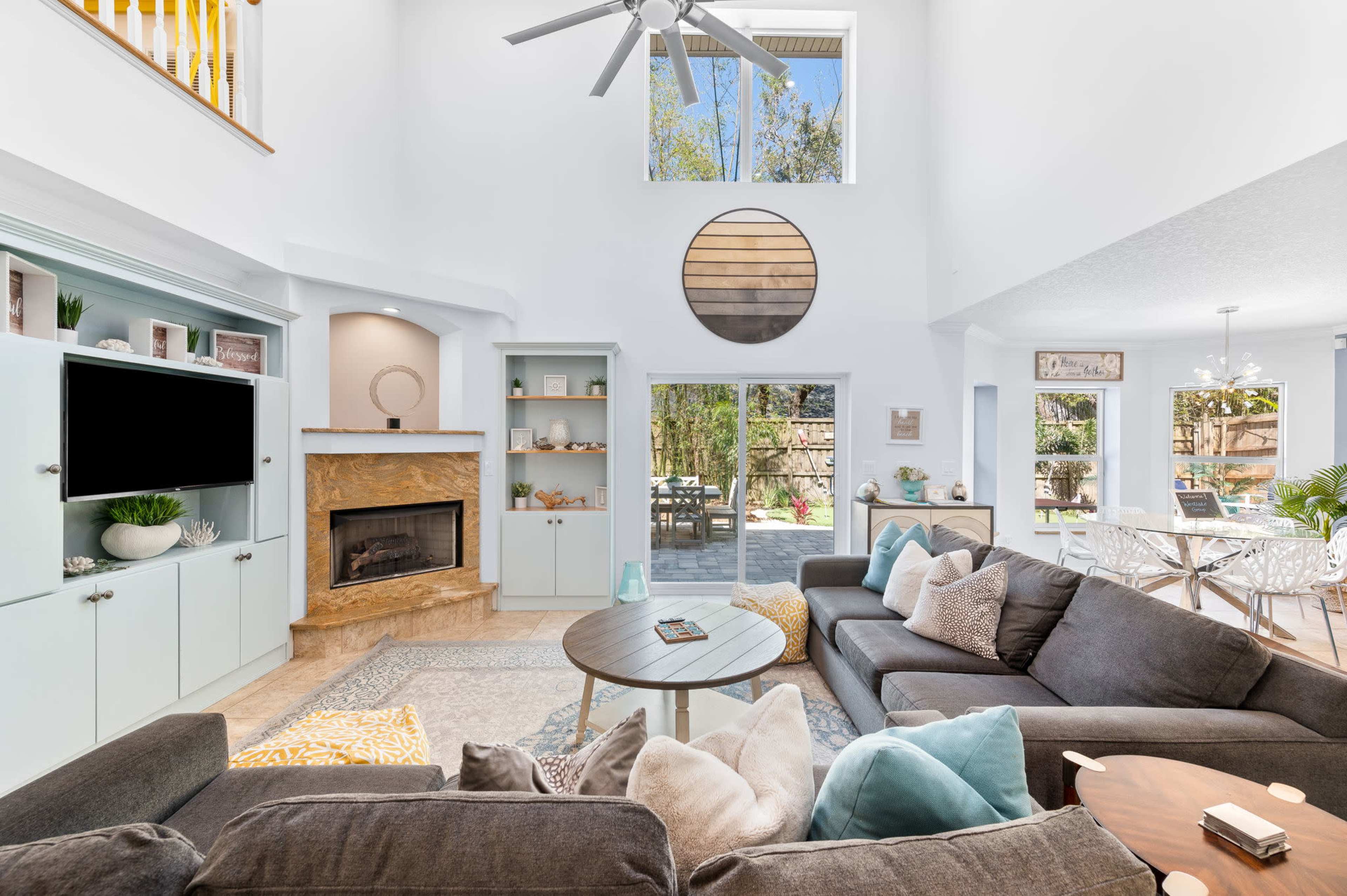 The image shows a bright, spacious living room with a high ceiling, featuring a large window, a stone fireplace, and comfortable seating arranged around a round coffee table.