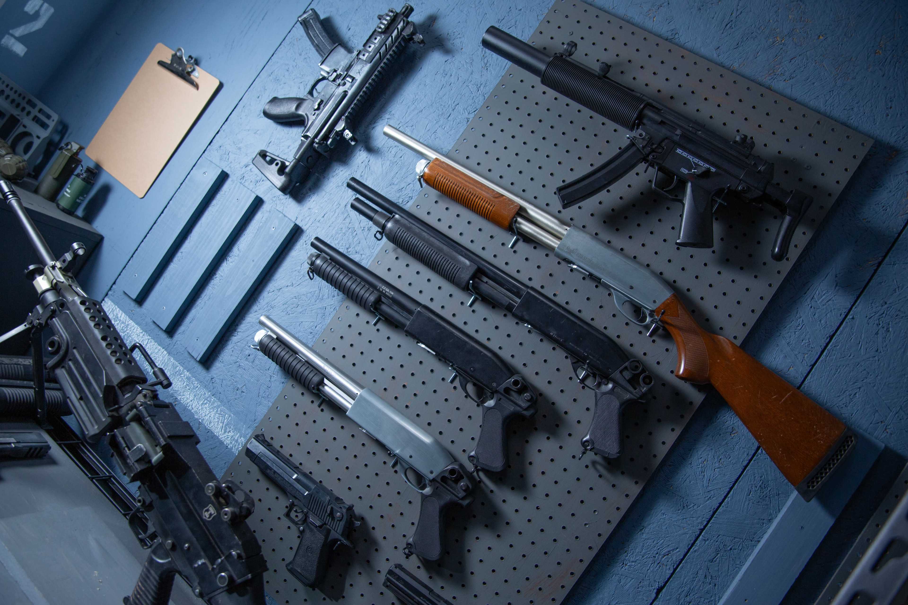 The image shows a variety of firearms displayed on a perforated panel against a blue wall.
