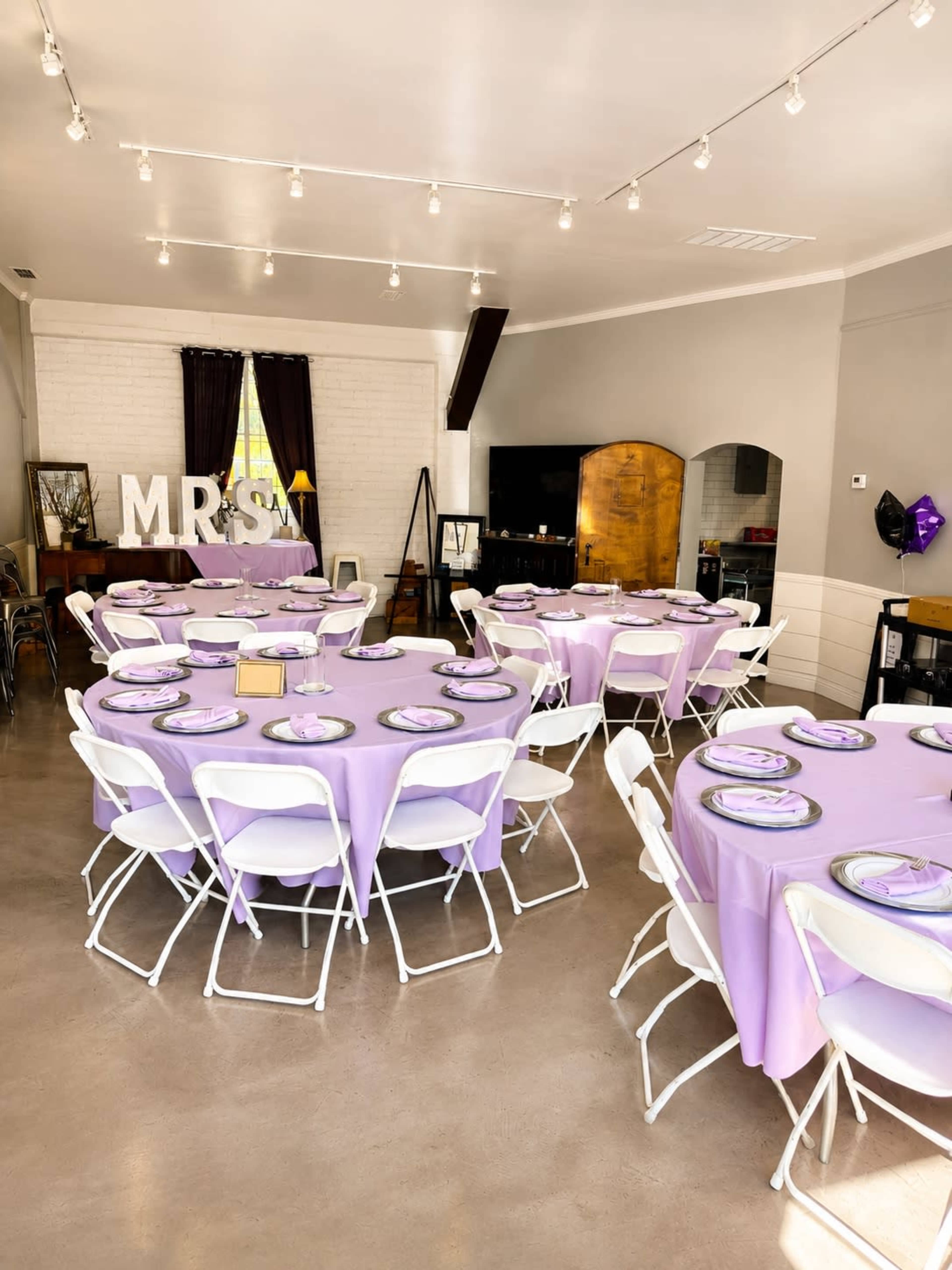 The image shows a decorated indoor event space with round tables covered in lavender cloth, each set with plates and chairs, and large letters spelling "MRS" on display.