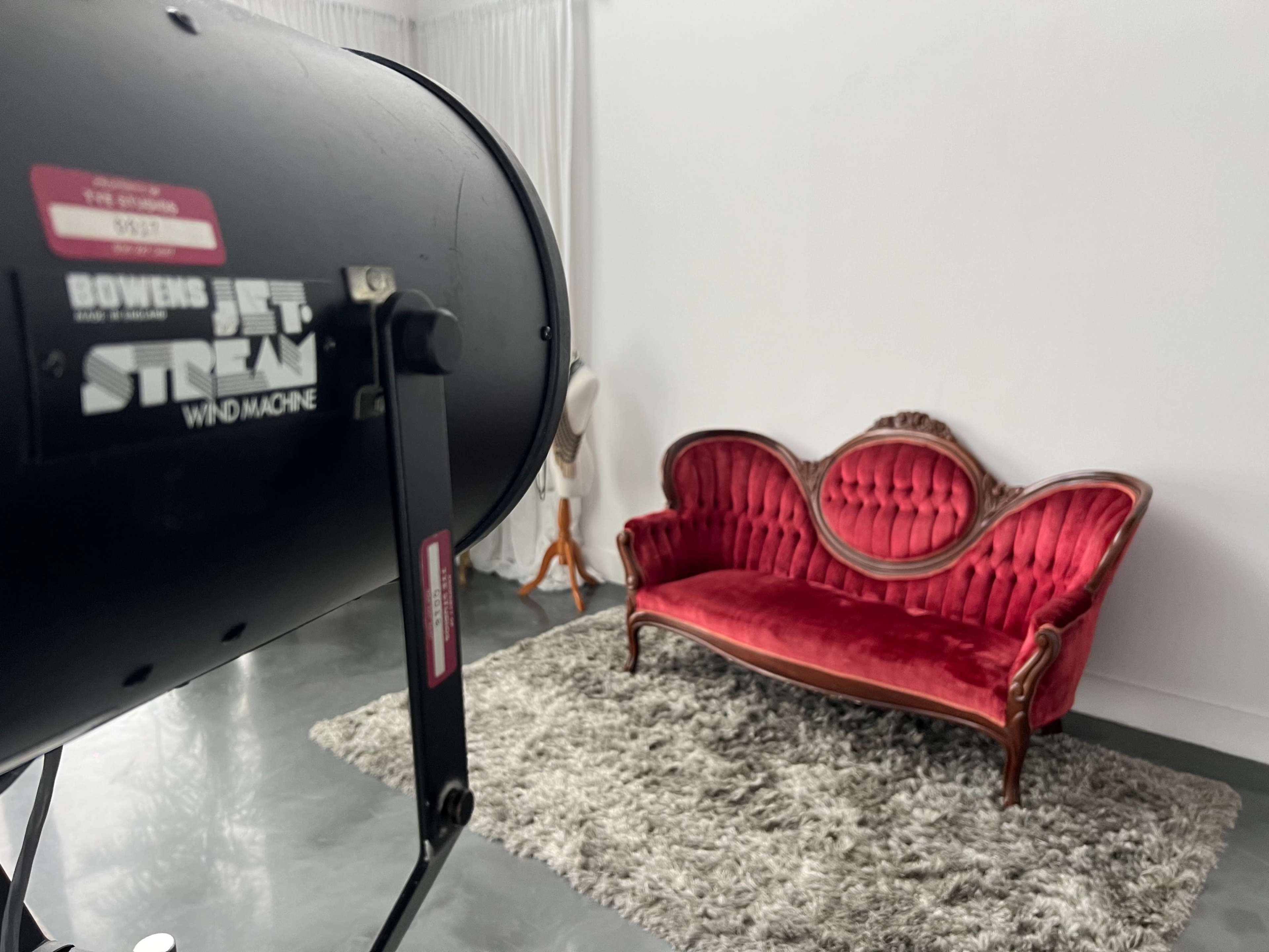 A vintage red velvet sofa is positioned on a plush gray rug in a bright studio space, with a spotlight aimed toward it.