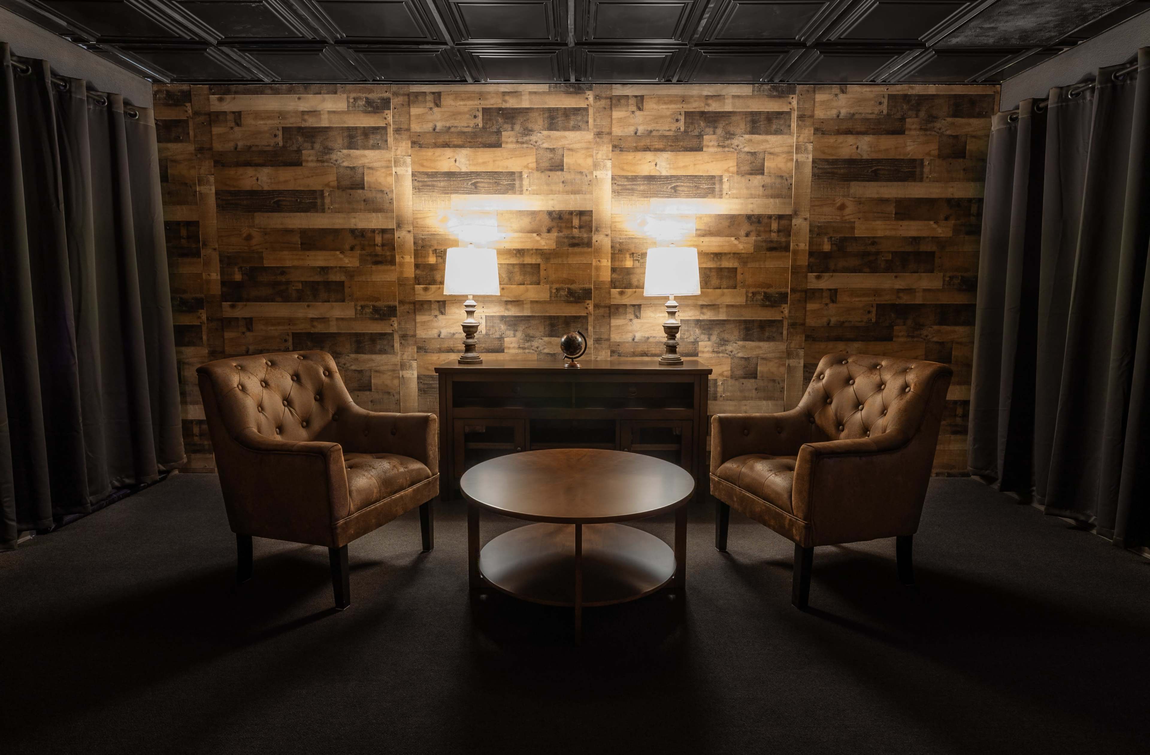 A cozy seating arrangement featuring two tufted leather chairs and a round coffee table, set against a wooden accent wall and illuminated by two table lamps.