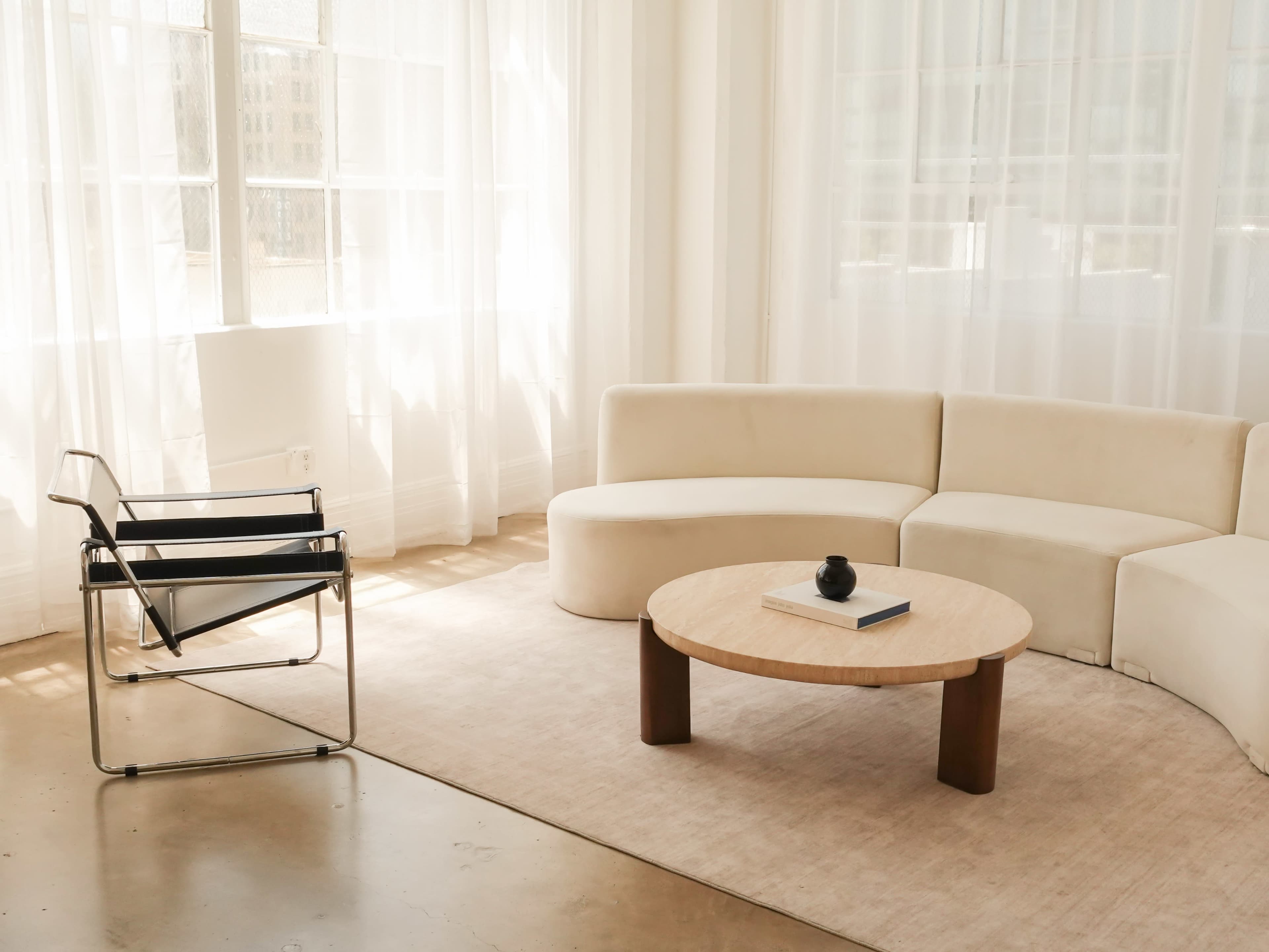 The image shows a minimalist living space featuring a curved beige sofa, a round wooden coffee table, and a black chair, with large windows covered by sheer curtains allowing natural light to filter in.