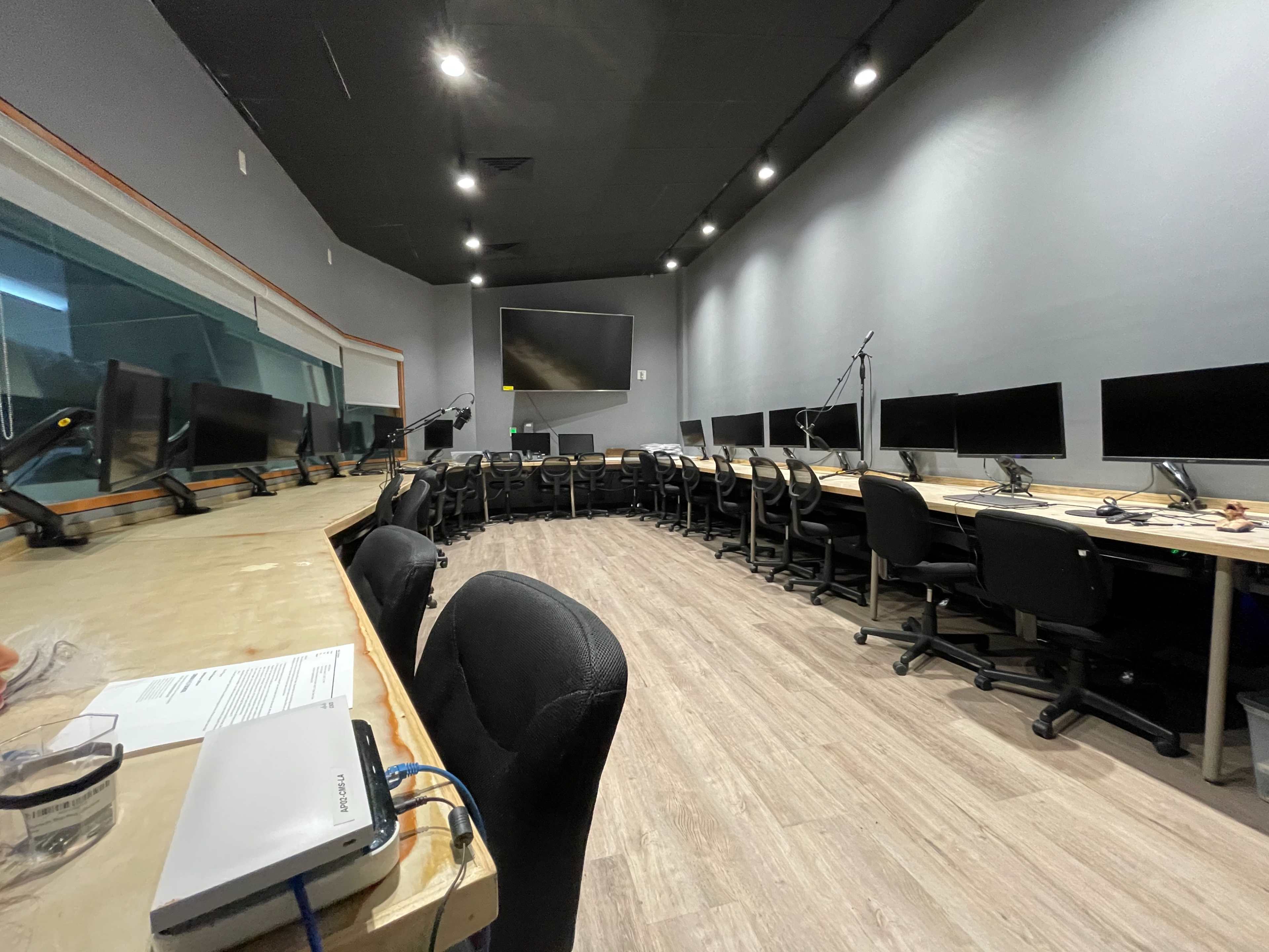 The image shows a modern recording studio with a circular arrangement of desks and monitors, equipped with chairs and a large screen at the front.