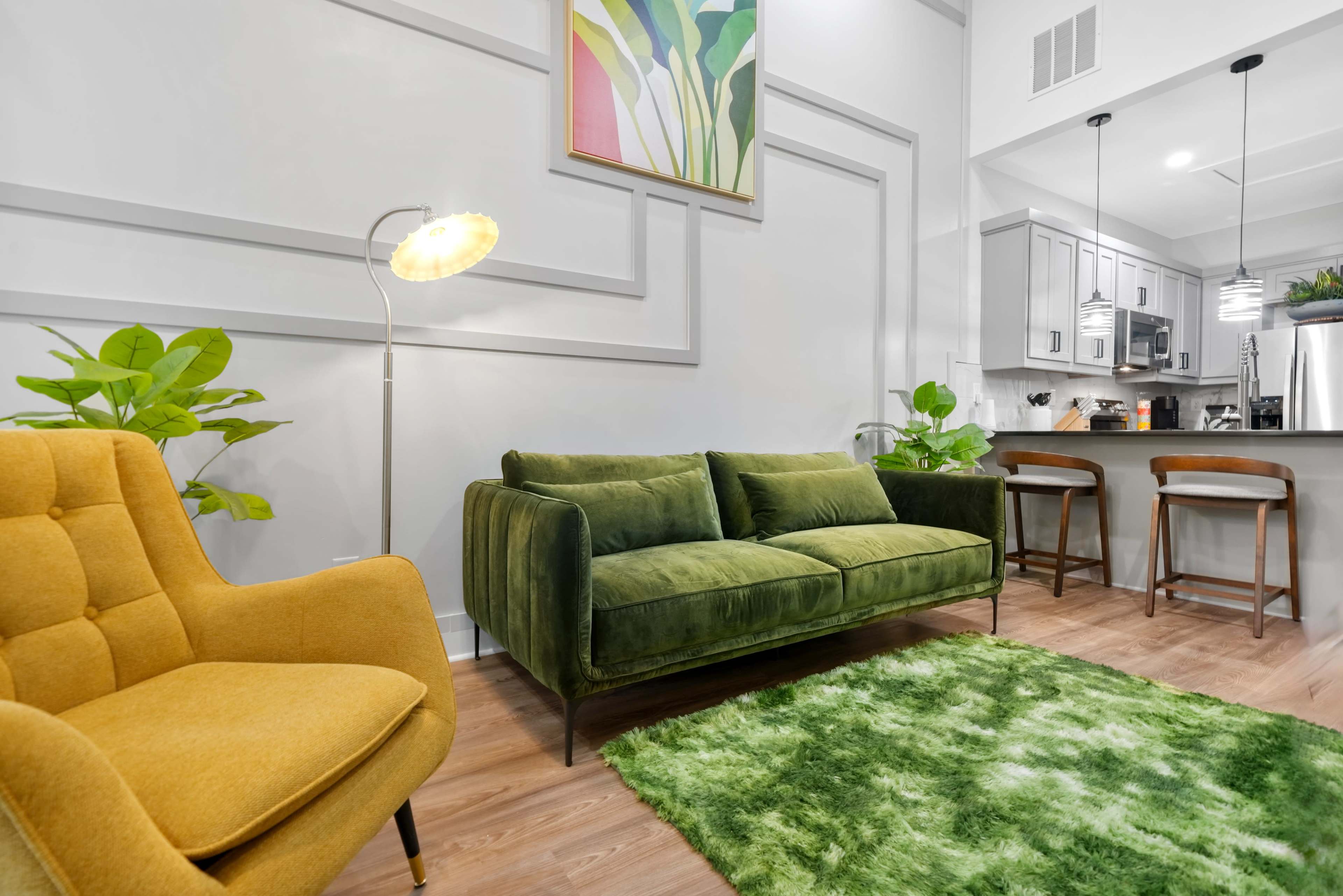 A modern living room features a green sofa and an orange armchair, with a patterned rug and plants, alongside a kitchen area in the background.
