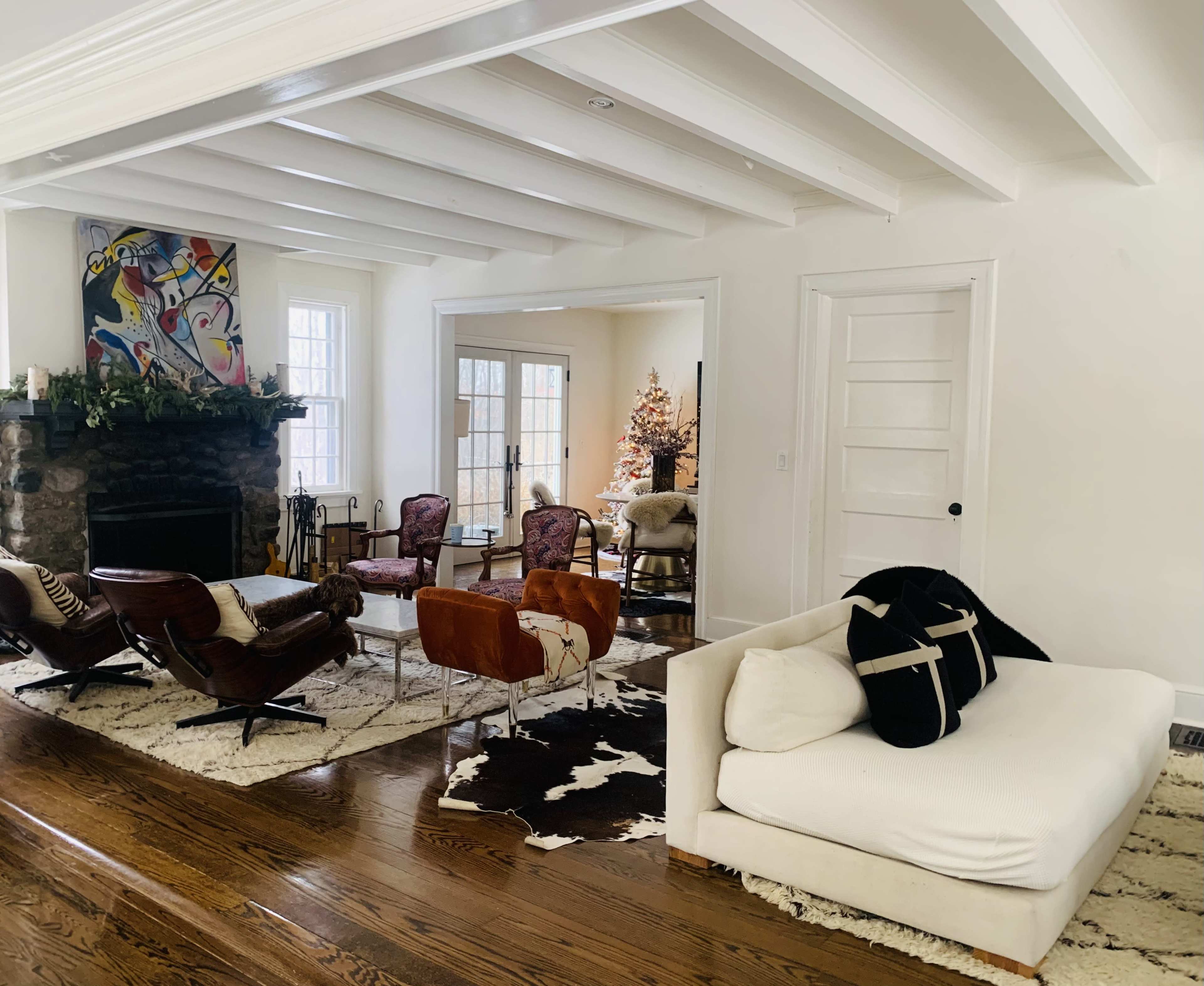 A bright living room featuring a white sofa, wooden flooring, and a stone fireplace, with a cozy arrangement of chairs and a view of a decorated tree in an adjoining room.
