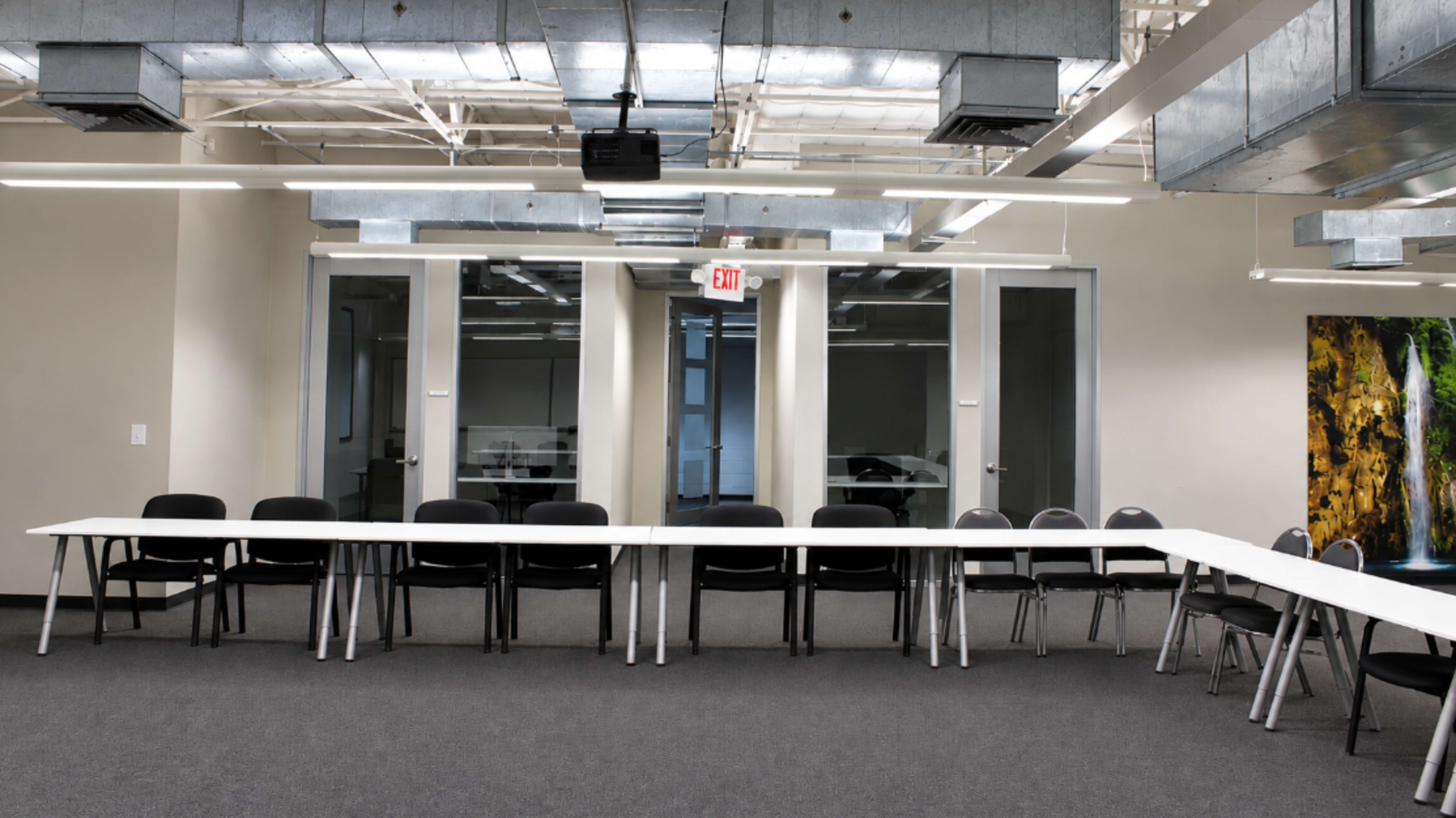 A long, white table is set up in a conference room with black chairs arranged around it, against a backdrop of glass doors and light-colored walls.