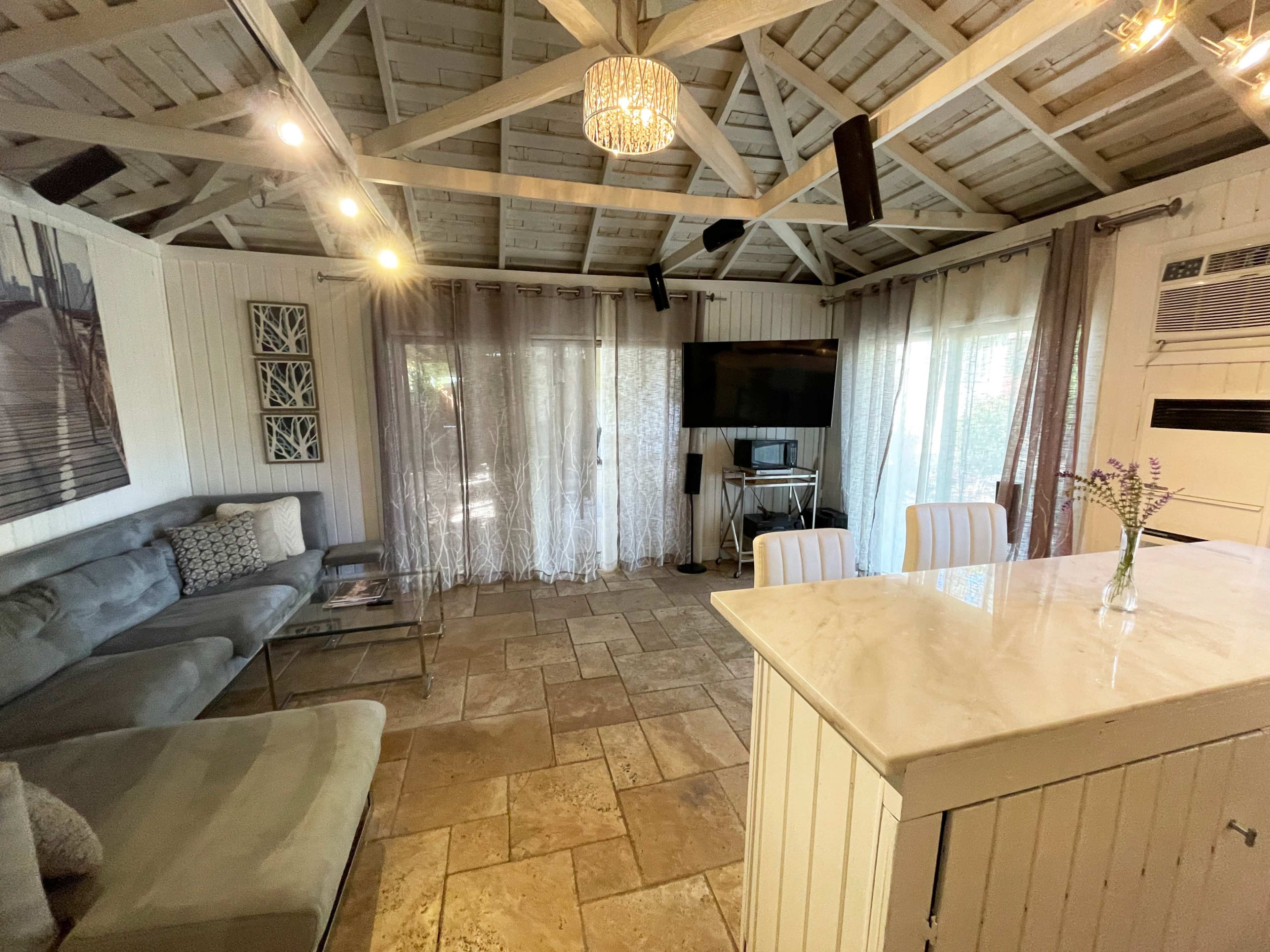 The interior of a cozy cottage features a stone floor, a large sectional sofa, a television against the wall, and a kitchen island with a light fixture above.