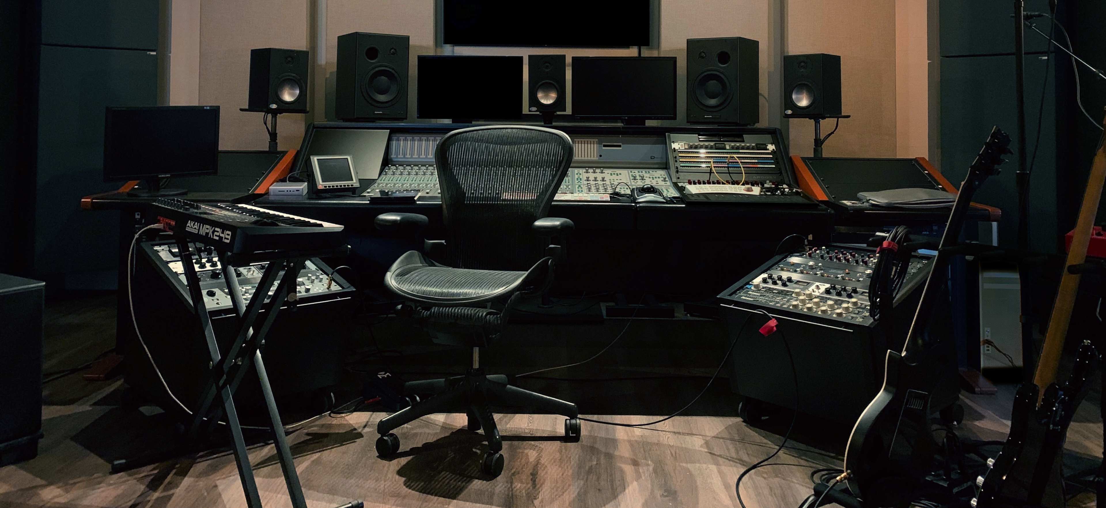 A music production studio with a central mixing console, multiple monitors, speakers, and various music equipment arranged around an ergonomic office chair.