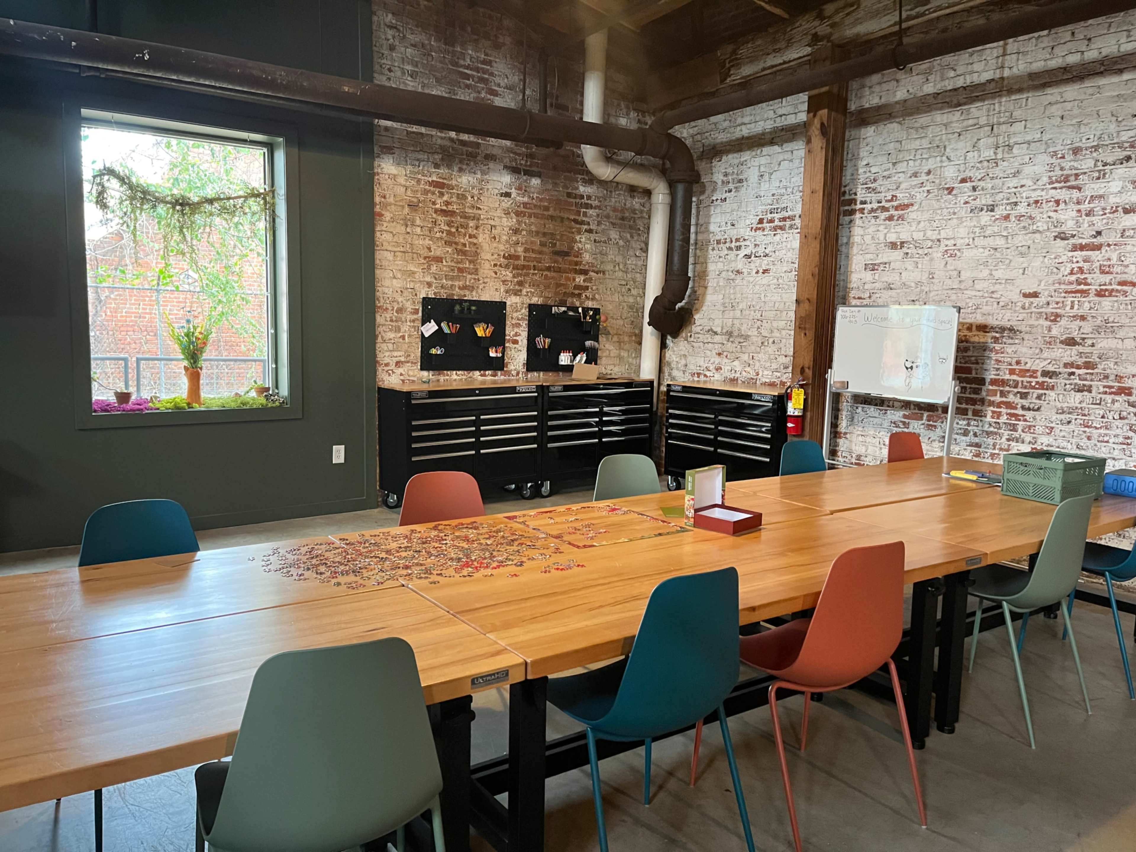The image shows a modern, open workspace with a large wooden table surrounded by colorful chairs, a puzzle spread across the table, and industrial-style brick walls.