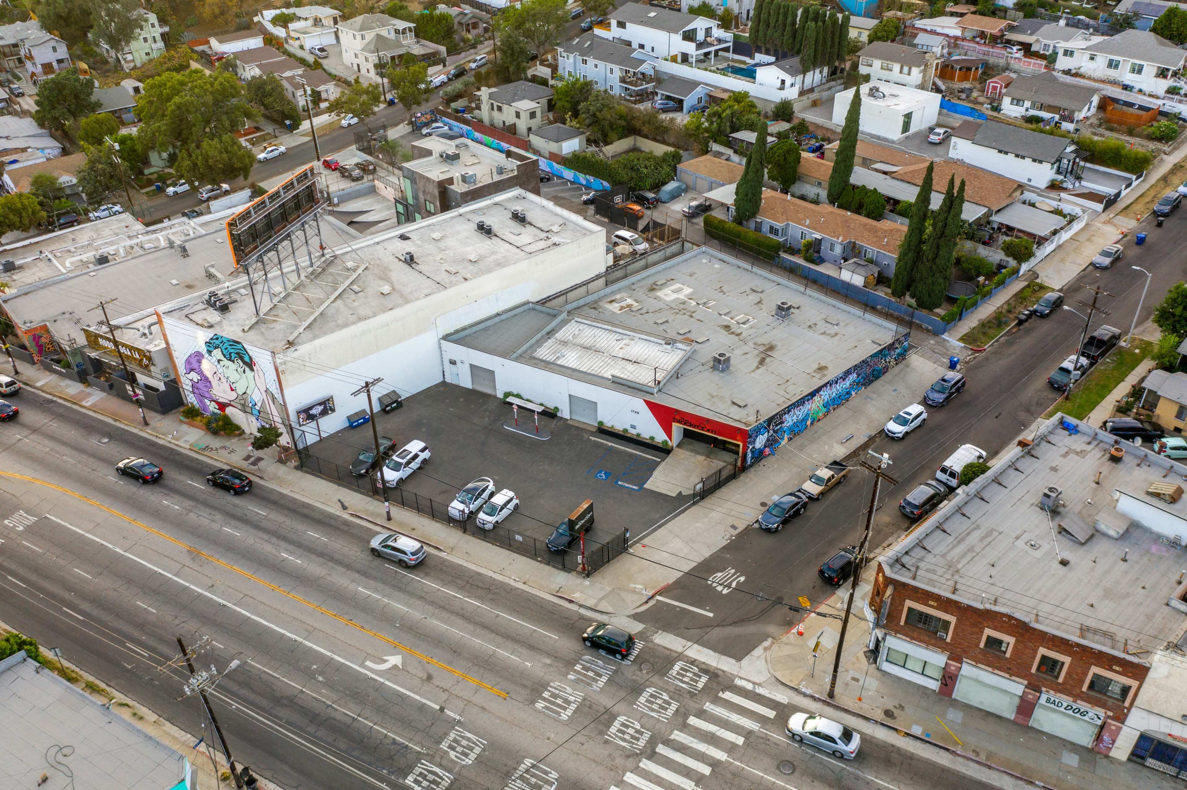 An aerial view of a commercial area featuring a large building with a mural, surrounded by several parked cars and nearby residential structures.
