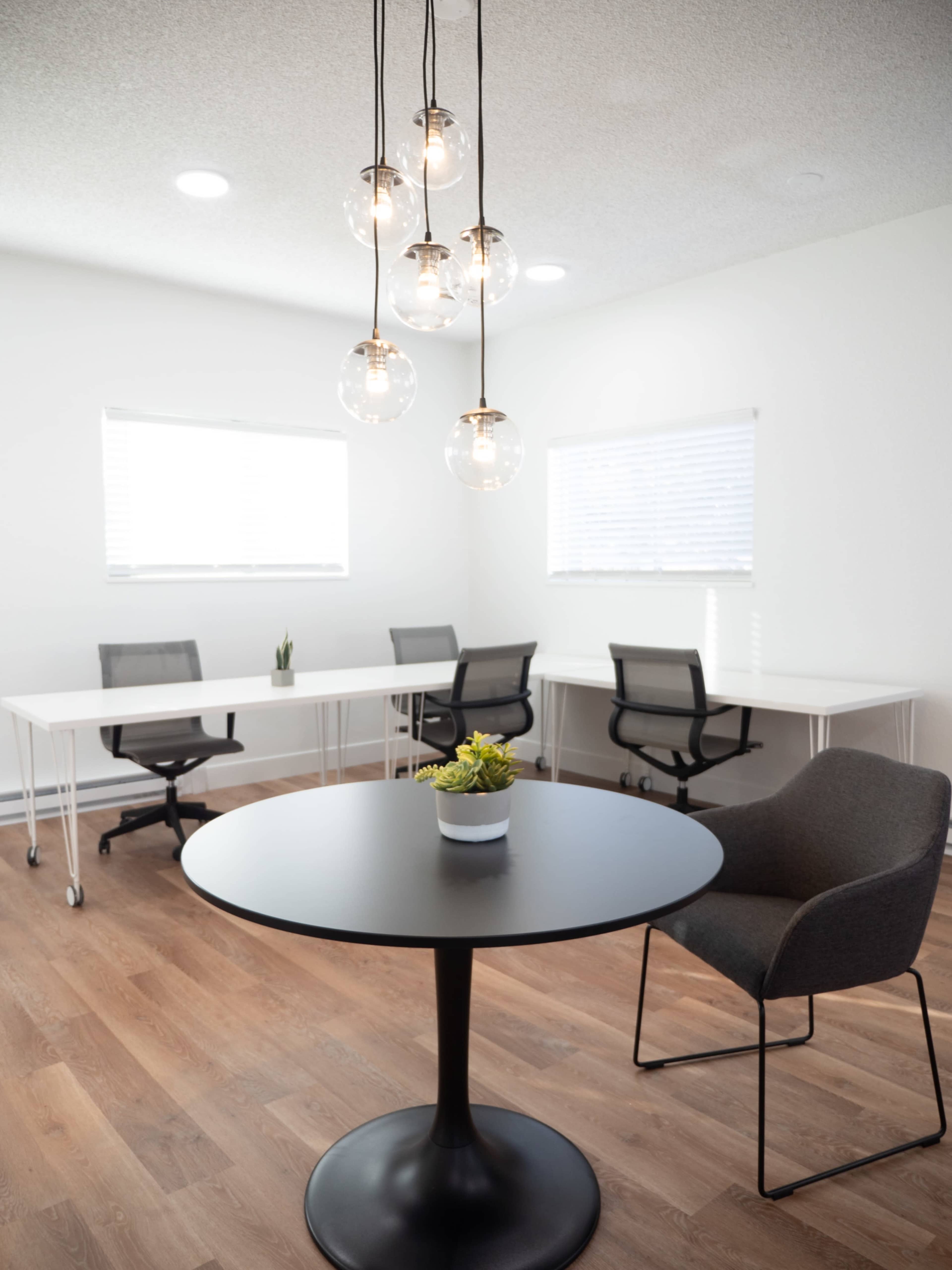 A modern office space features a round table with a plant in the center and four desks with rolling chairs in the background, illuminated by pendant lighting.