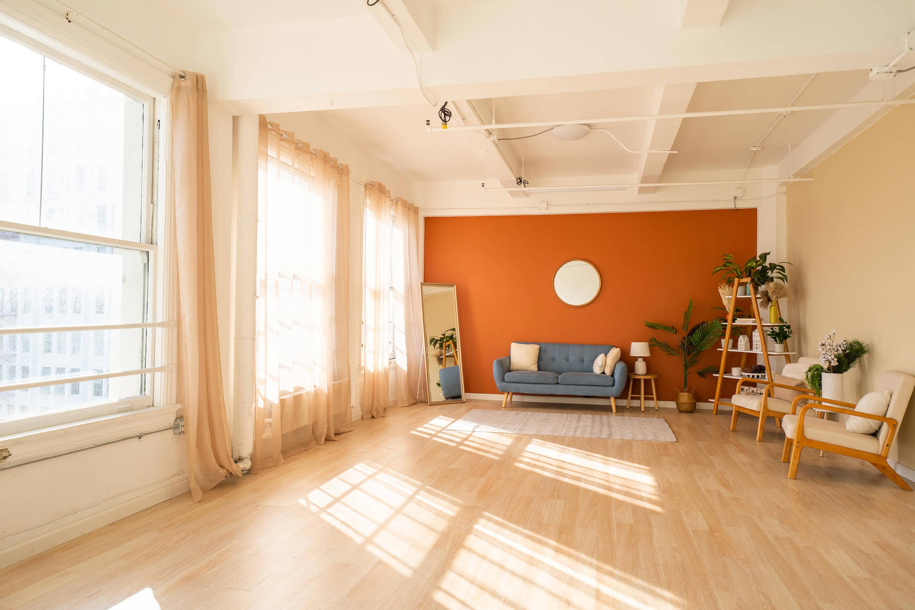 The image shows a bright, spacious room featuring a blue sofa, a ladder bookshelf, and large windows with sheer curtains, all set against an orange wall.