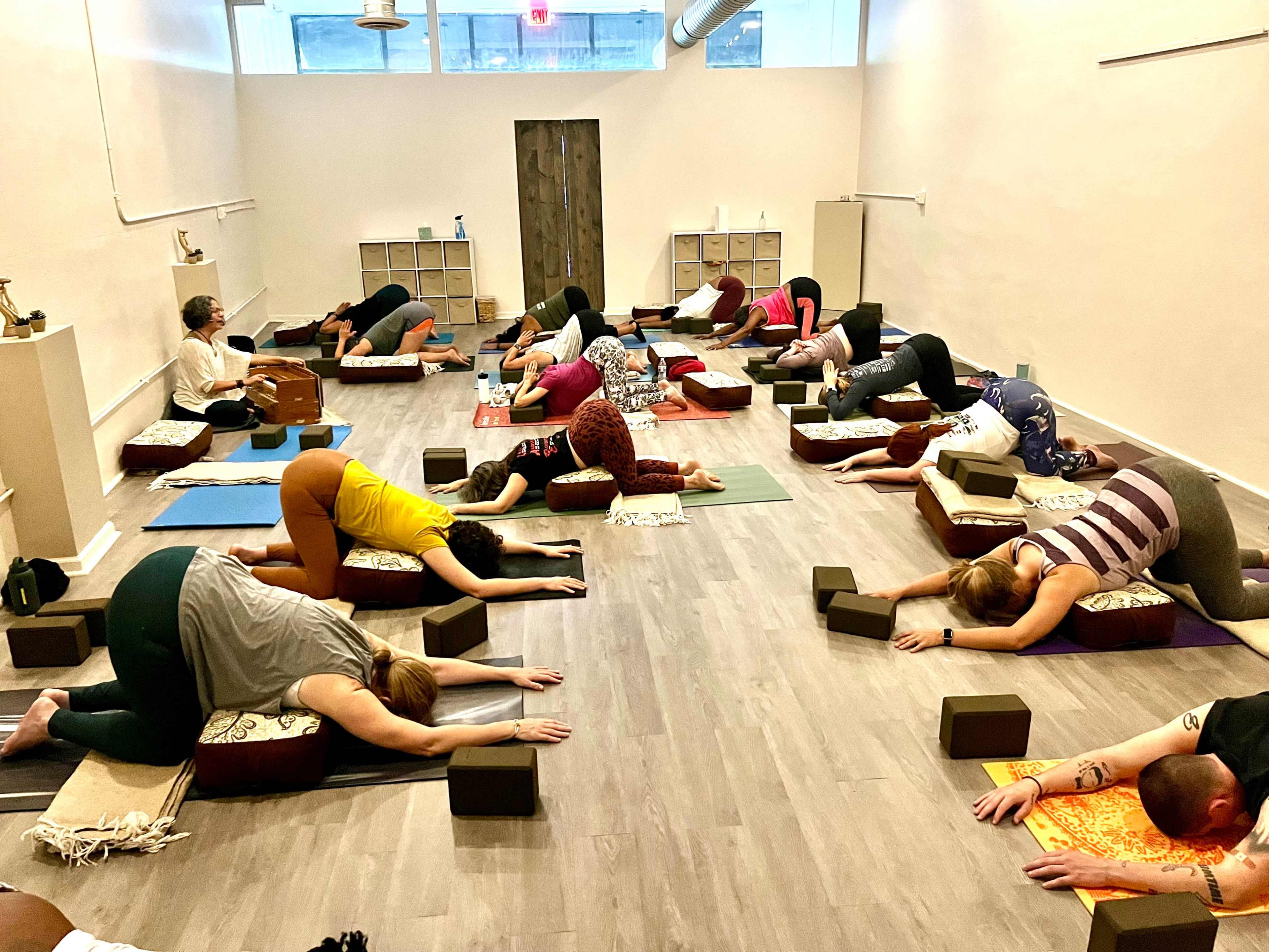 A group of people is participating in a yoga class in a spacious, well-lit studio, positioned on mats and cushions in various relaxed postures.
