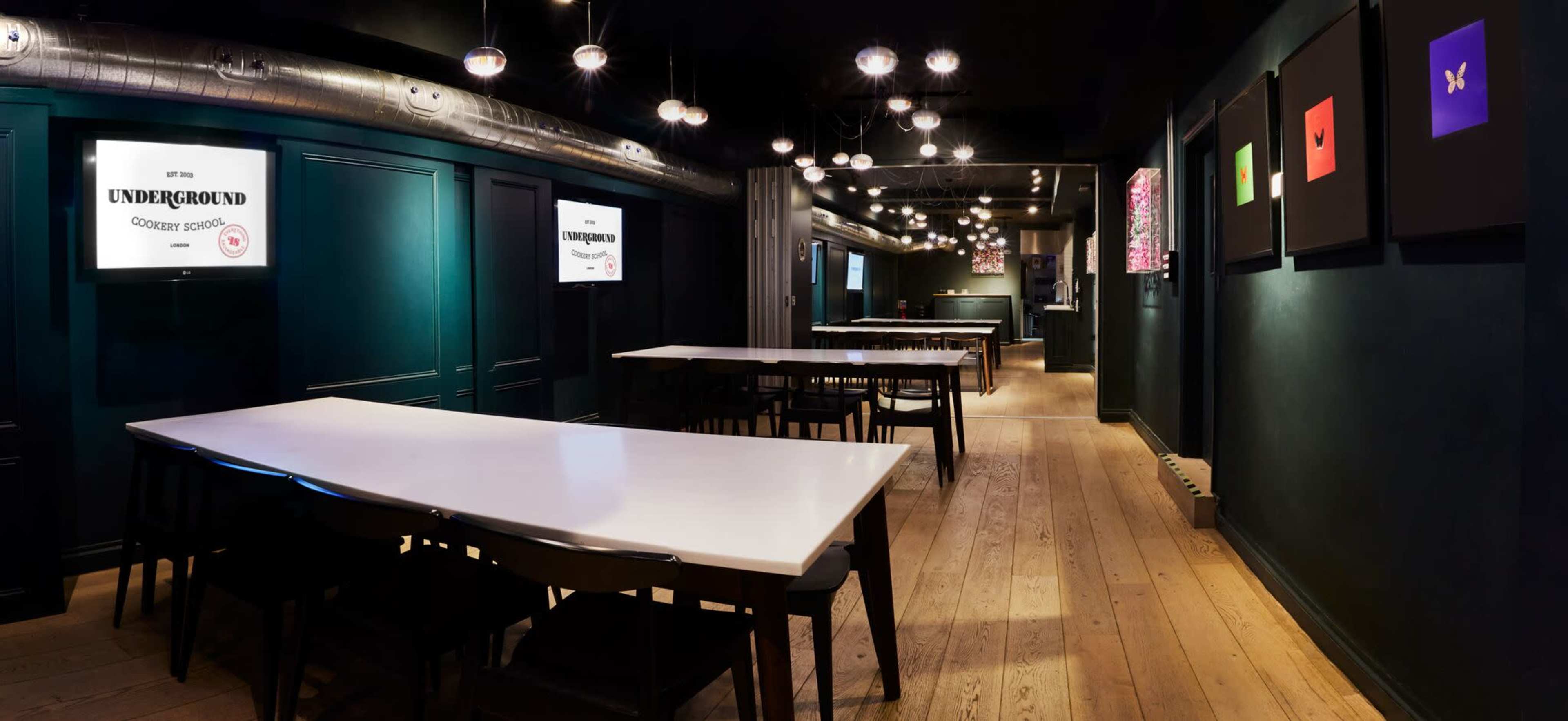 The image shows a dimly lit restaurant interior featuring long white tables, black chairs, and framed colorful artwork on dark walls, with hanging light fixtures overhead.