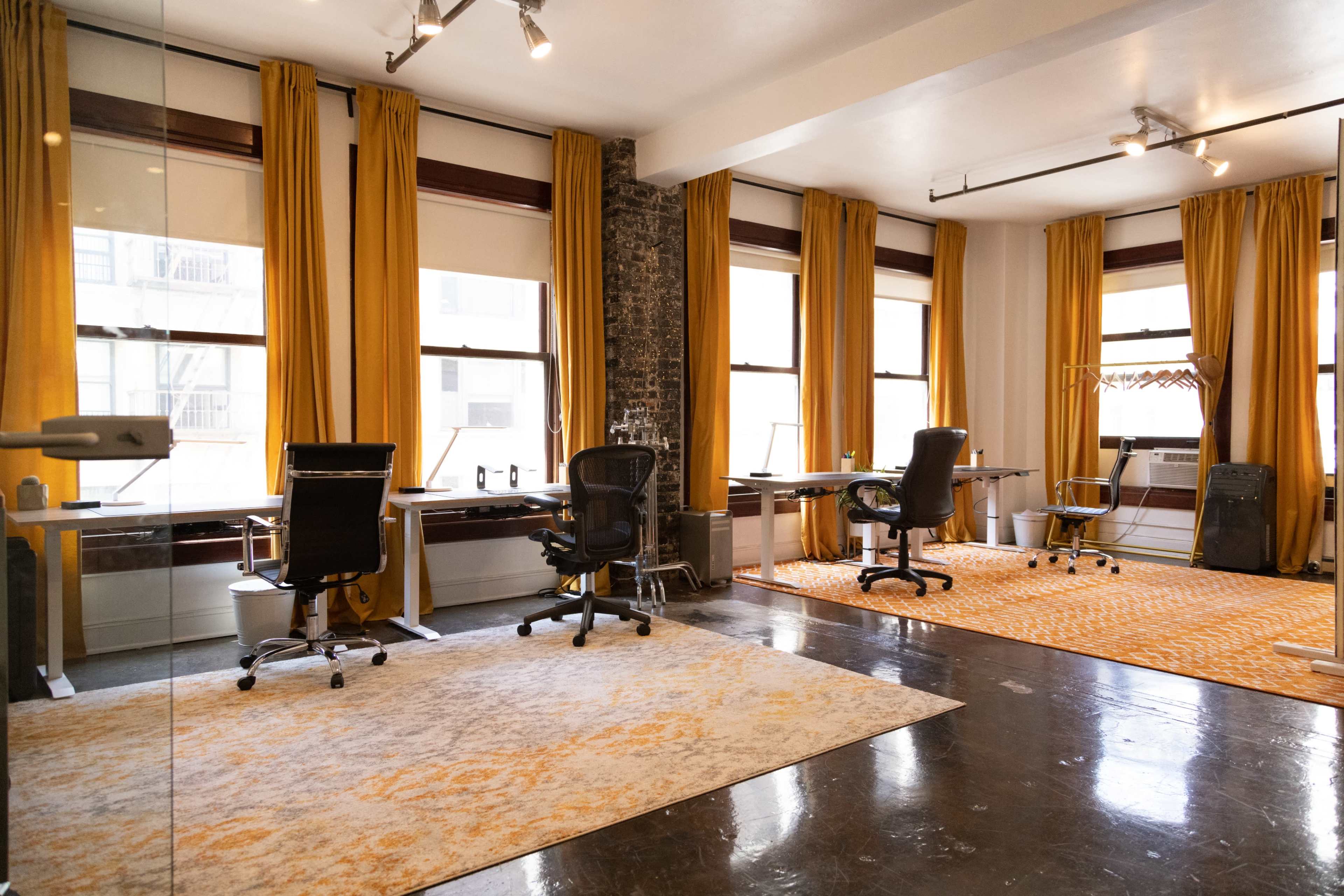 The image shows a modern office space with two desks, ergonomic chairs, and large windows featuring yellow curtains, set against a backdrop of exposed brick and a patterned area rug.