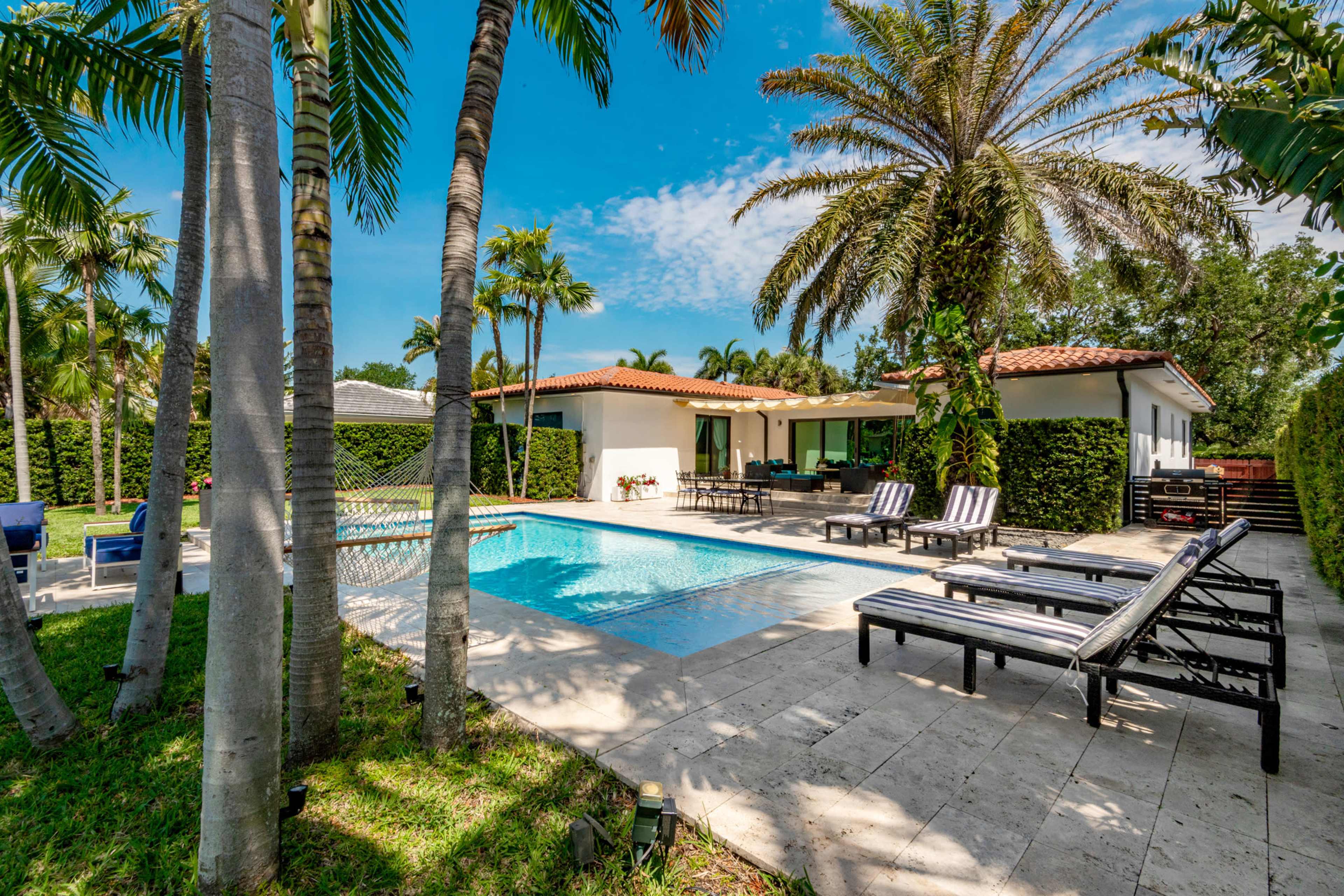 The image shows a landscaped outdoor area featuring a swimming pool, lounge chairs, and tropical palm trees under a clear blue sky.