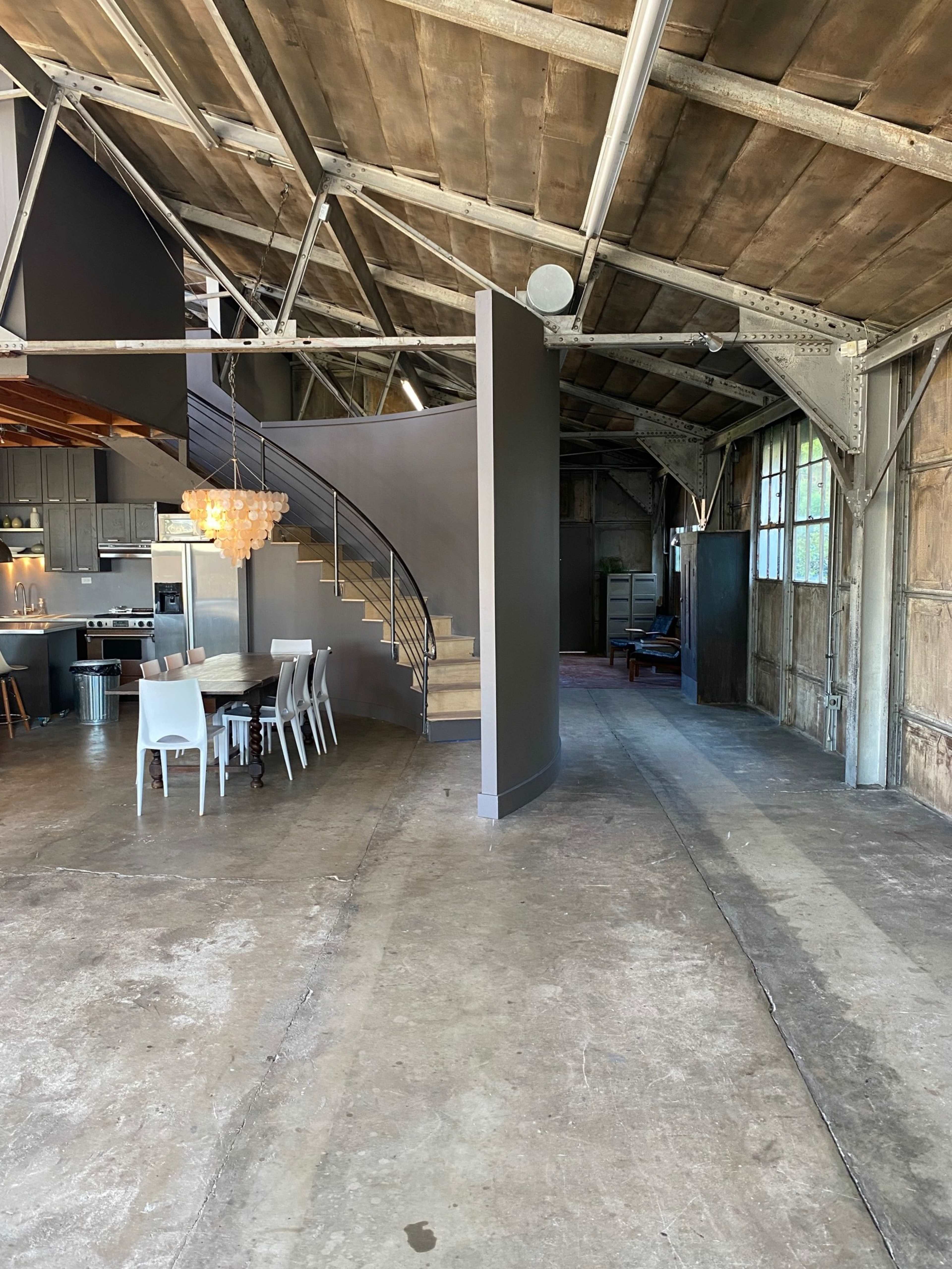 The image shows a spacious industrial-style interior with a curved staircase, a dining area with a round table and white chairs, and exposed beams in the ceiling.