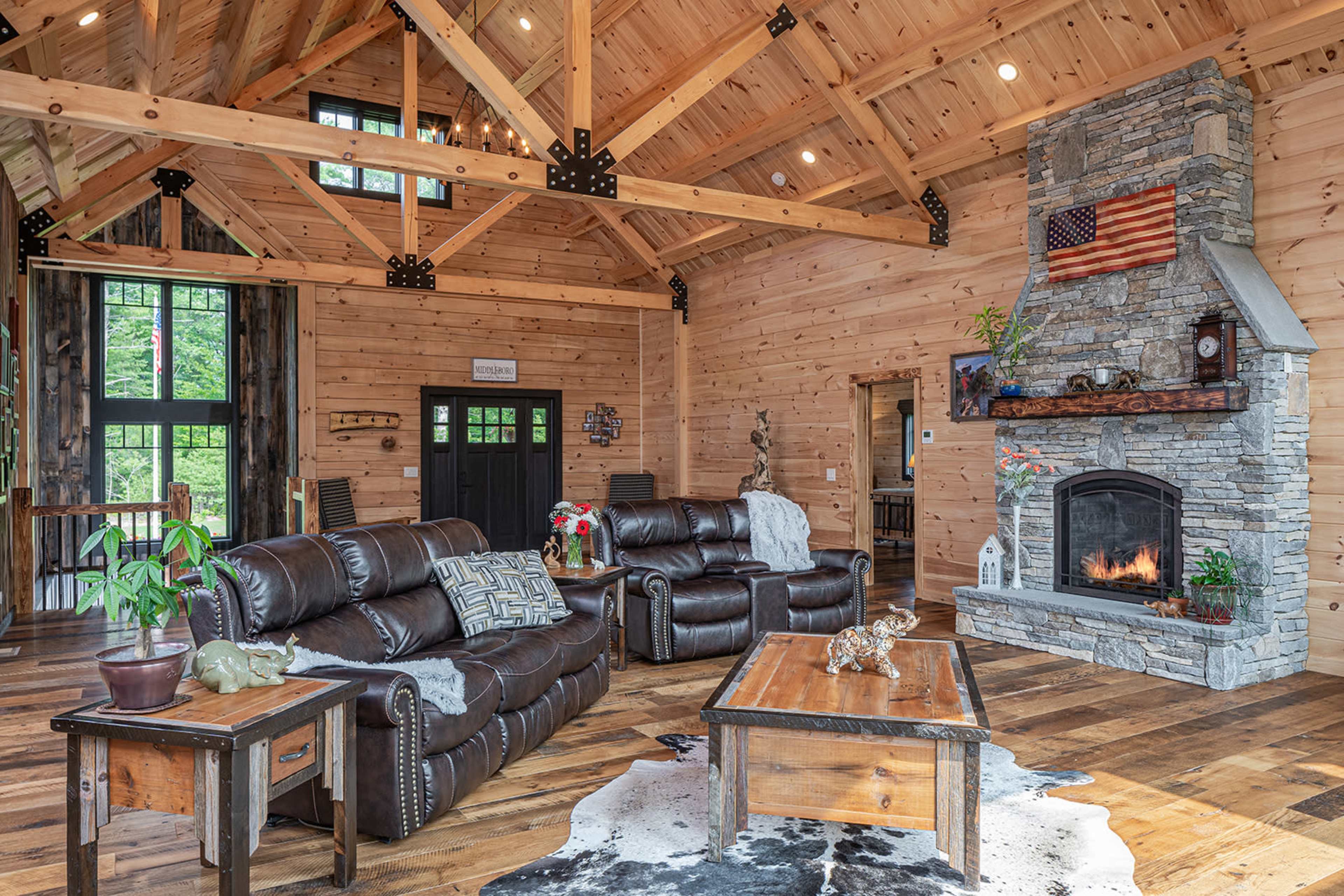 A spacious living room featuring wooden beams, a stone fireplace, and leather seating arranged around a central coffee table on a wooden floor.