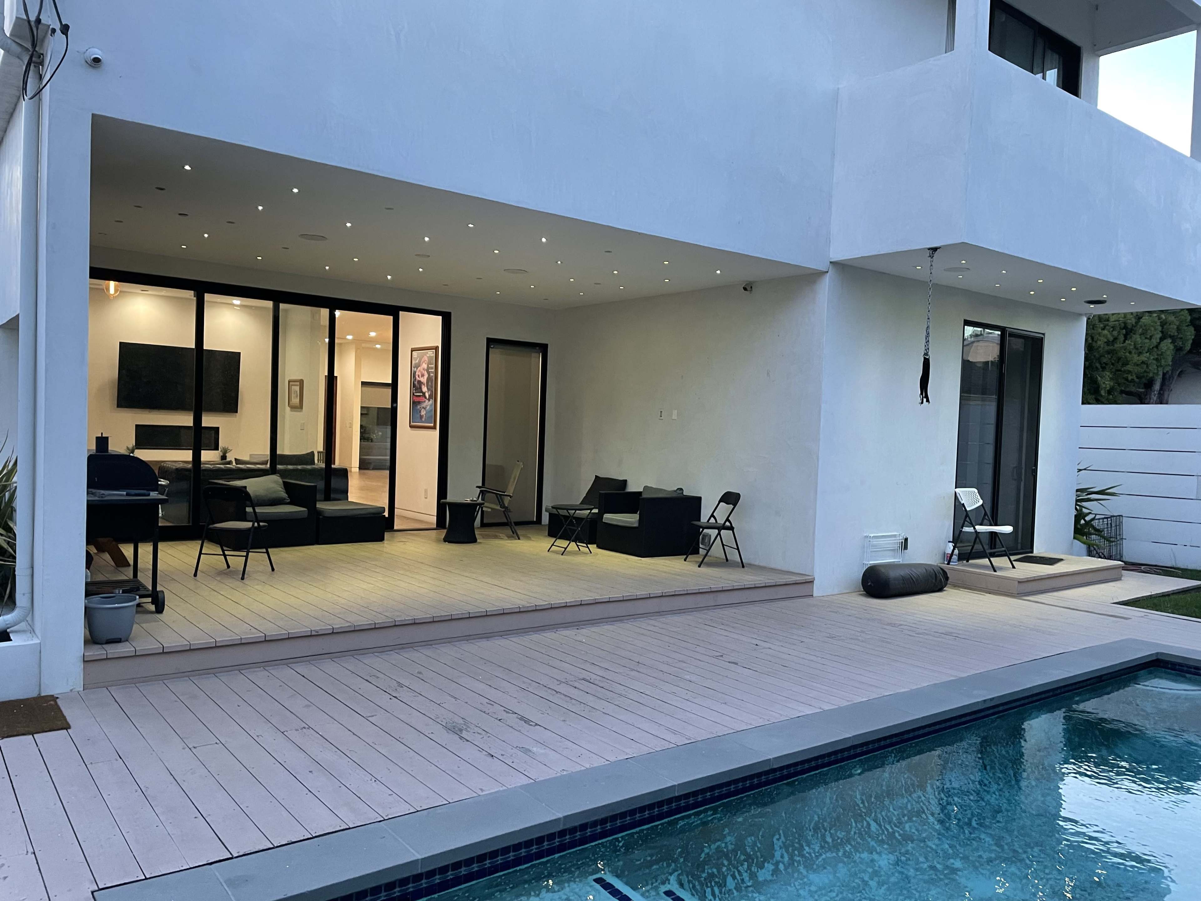 A modern two-story house features a pool, a wooden deck with chairs, and large glass doors leading into a living space.