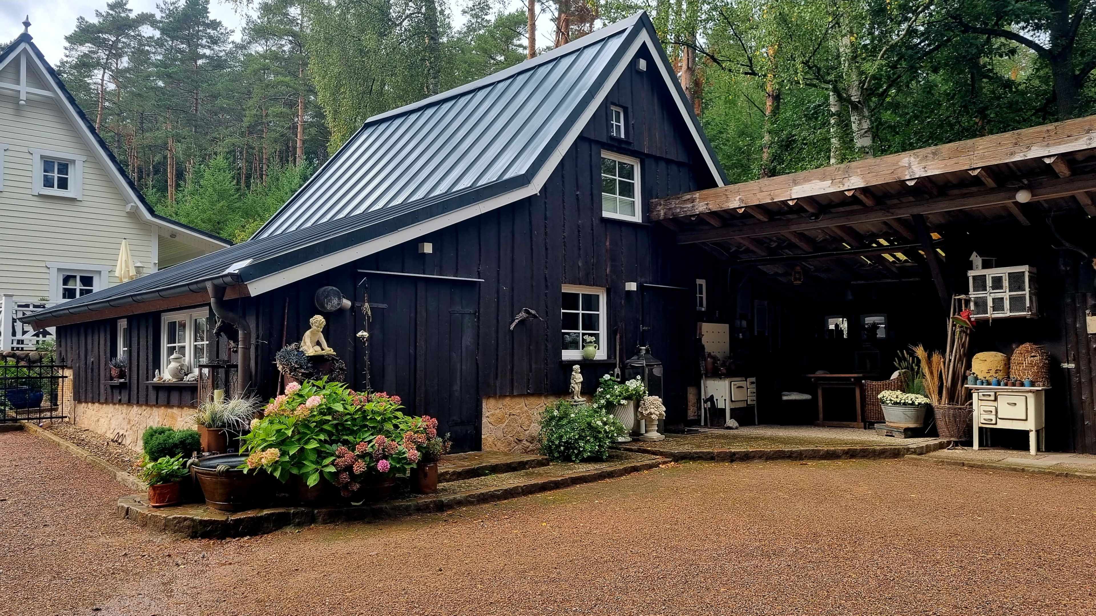 The image shows a black wooden cottage with a sloped roof, surrounded by greenery and various decorative plants, alongside a covered area that appears to serve as an outdoor workspace or storage.
