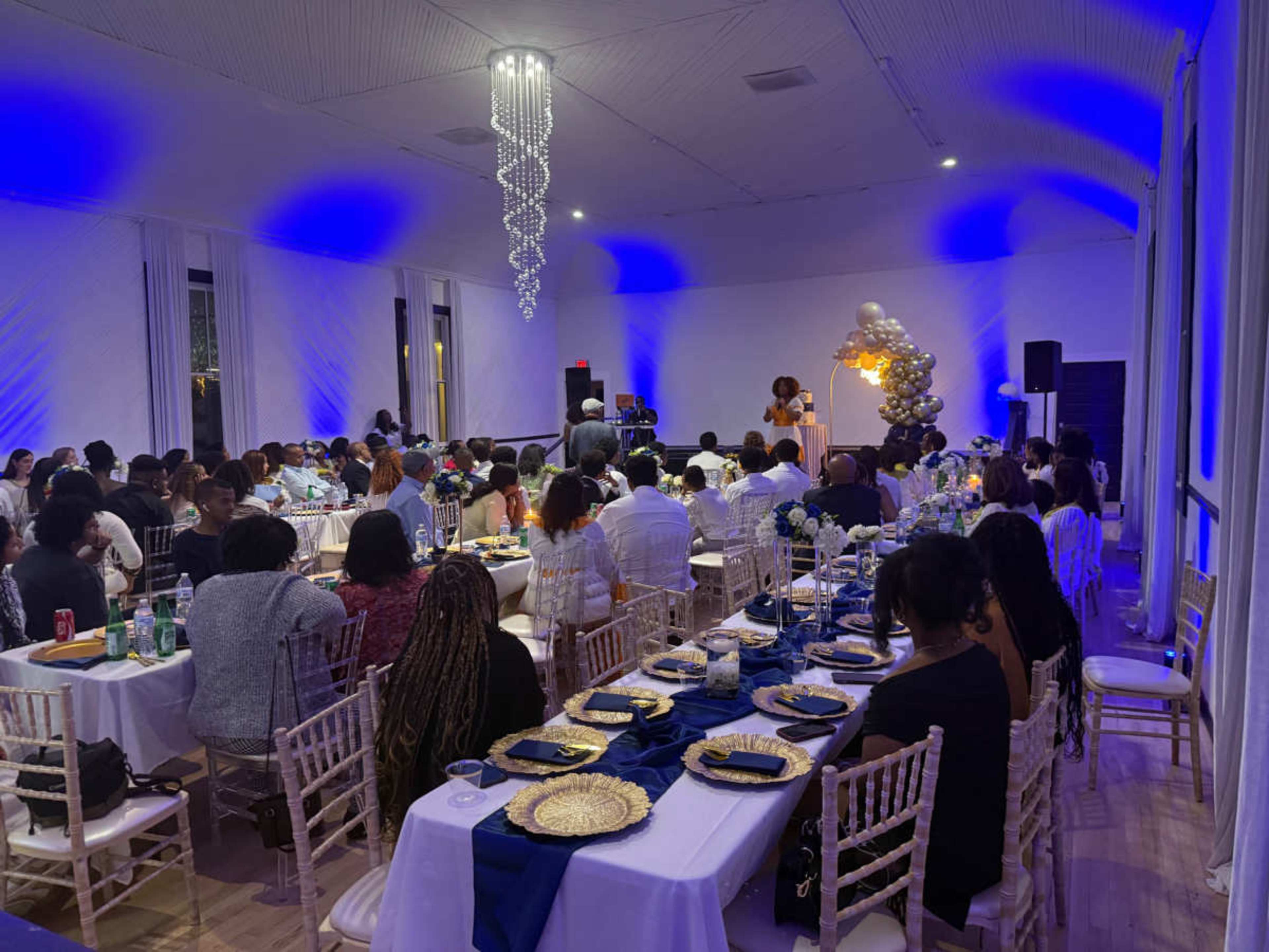 The image shows a large indoor event space filled with people seated at tables decorated with blue and gold tablecloths, while a speaker is addressing the audience from a small stage.