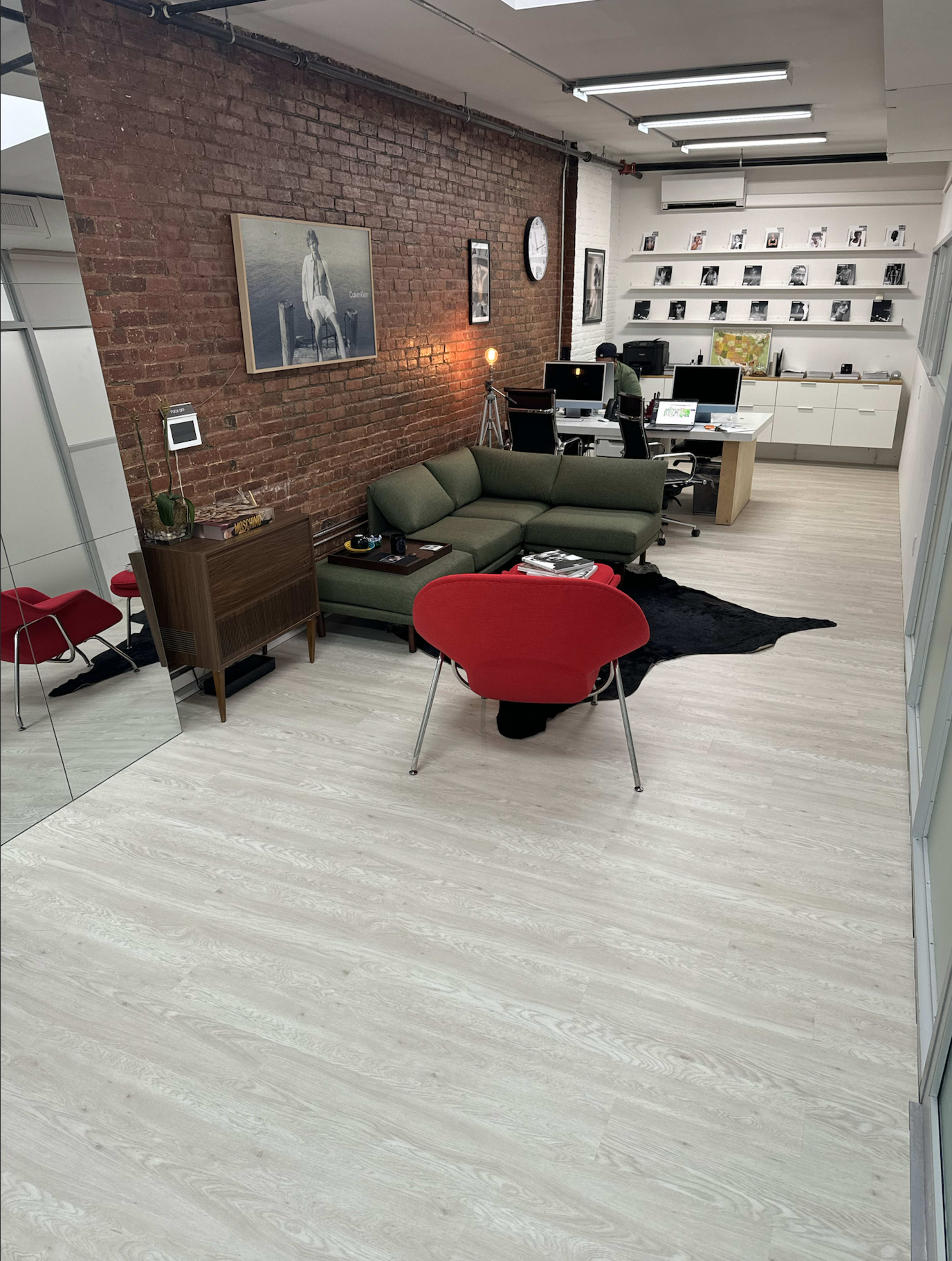The image shows a modern office space with a seating area featuring a green sofa and red chairs, alongside desks with computers and a bookshelf against a brick wall.