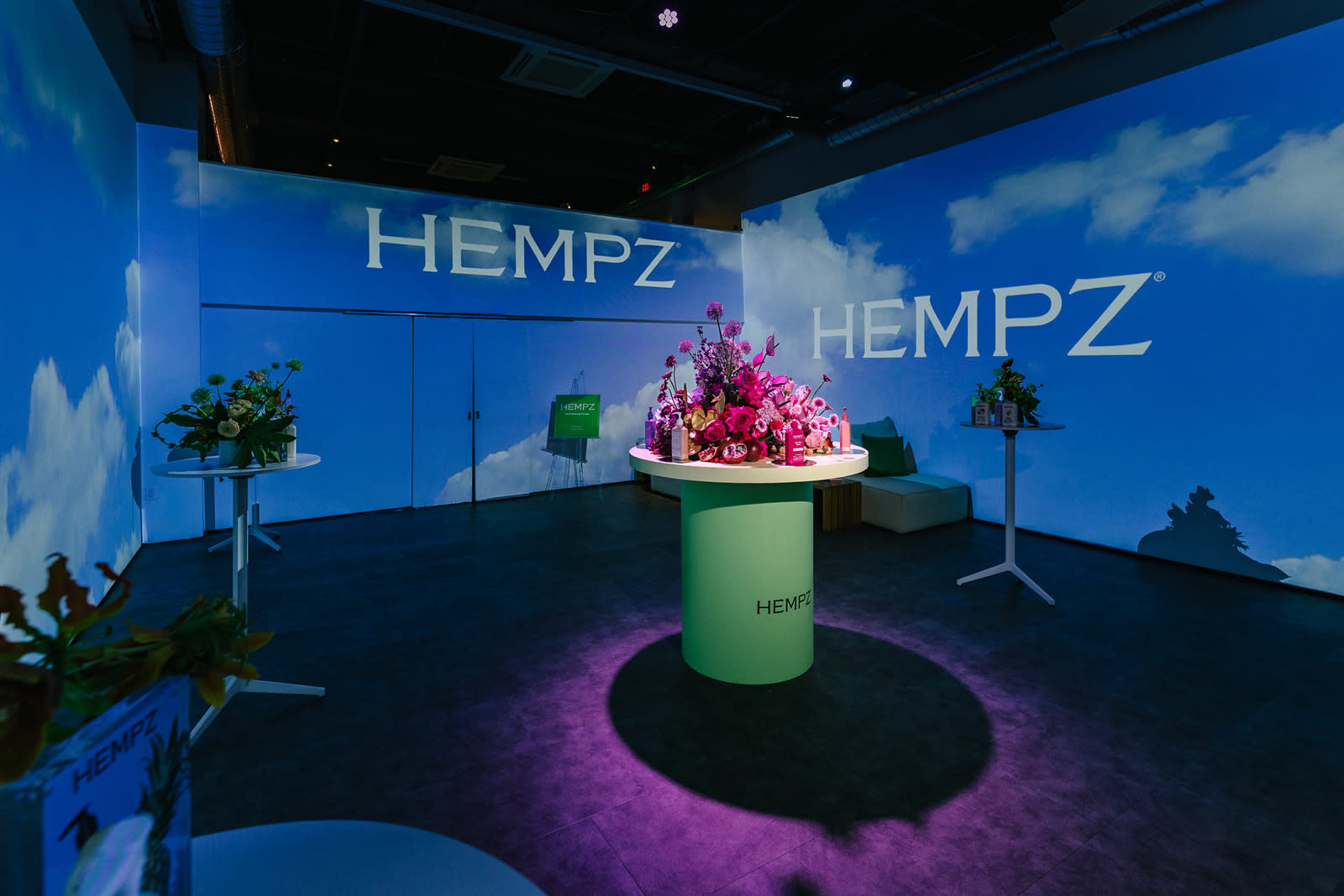 The image shows a modern indoor space with large promotional displays featuring the brand name "HEMPZ" on the walls, illuminated by soft lighting and decorated with floral arrangements on circular tables.