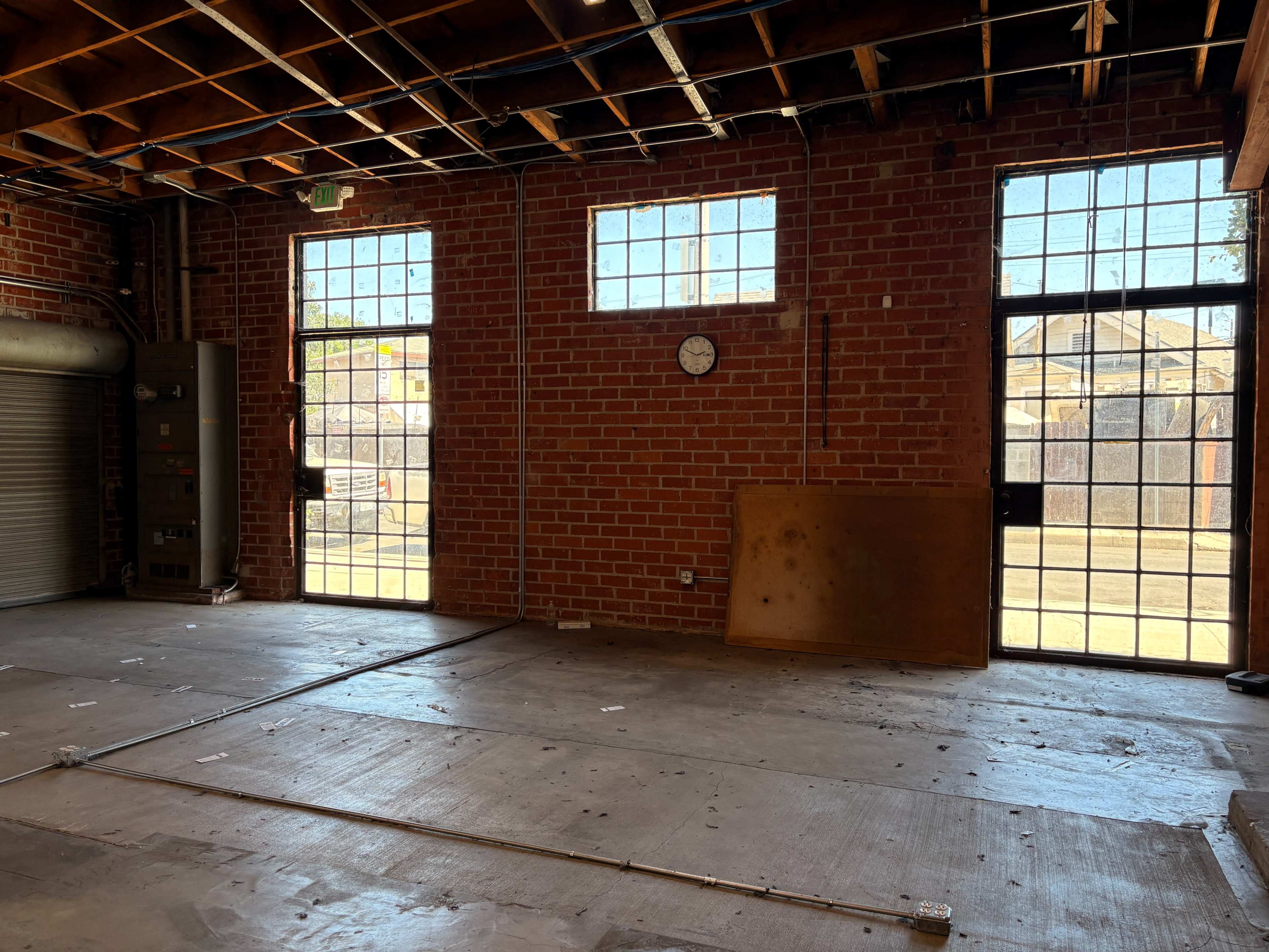 The image shows an empty brick room with large windows and a clock on the wall, featuring a wooden panel leaning against the floor.