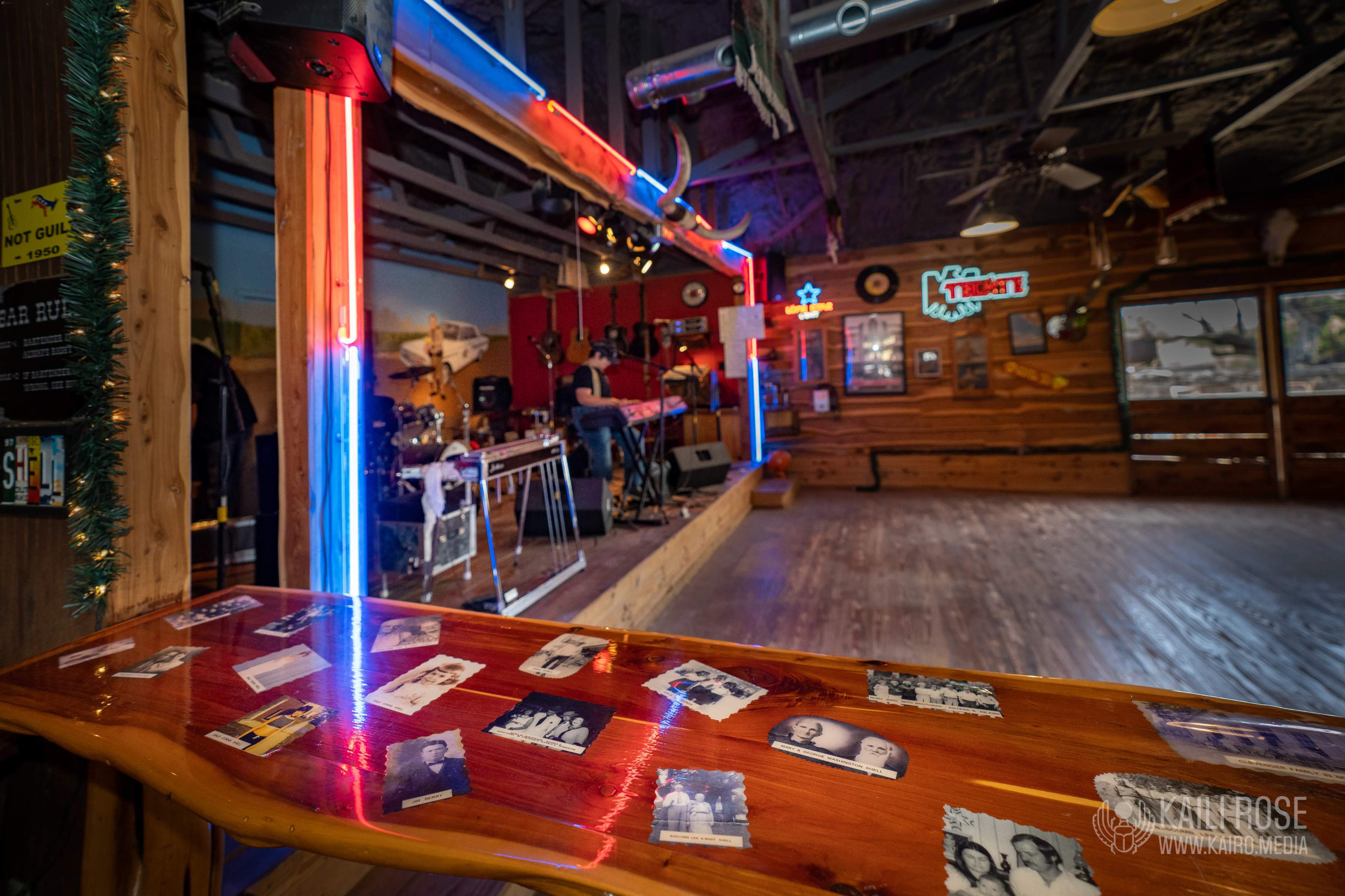 A stage is set up with musical instruments in a rustic wooden venue, featuring a bar adorned with black-and-white photographs.