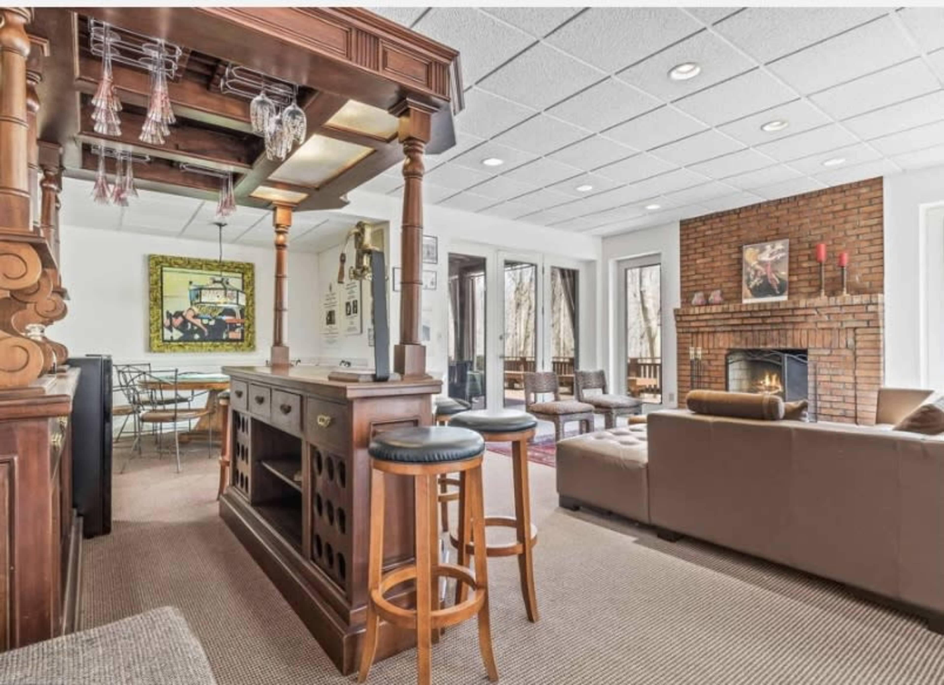 The image depicts a living space with a wooden bar area, seating, a fireplace, and large windows allowing natural light.