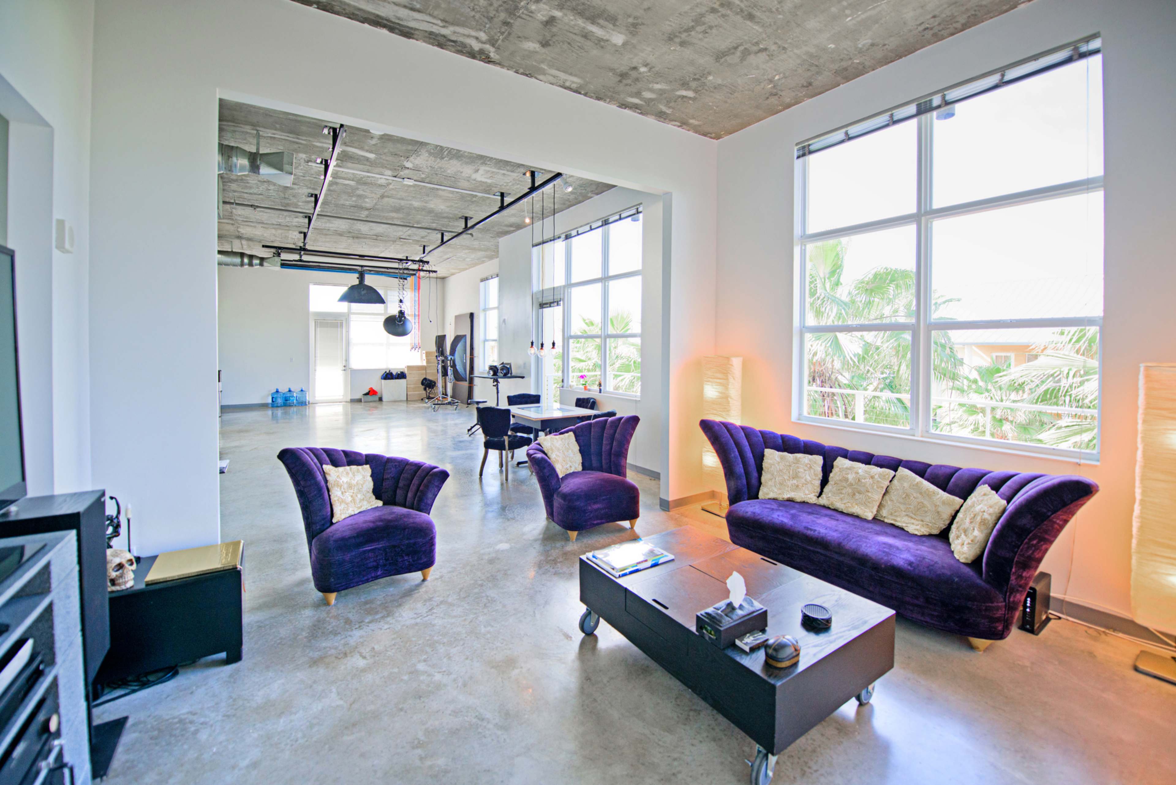 A spacious, modern living area features a purple velvet sofa set, a coffee table, and large windows allowing natural light to enter.