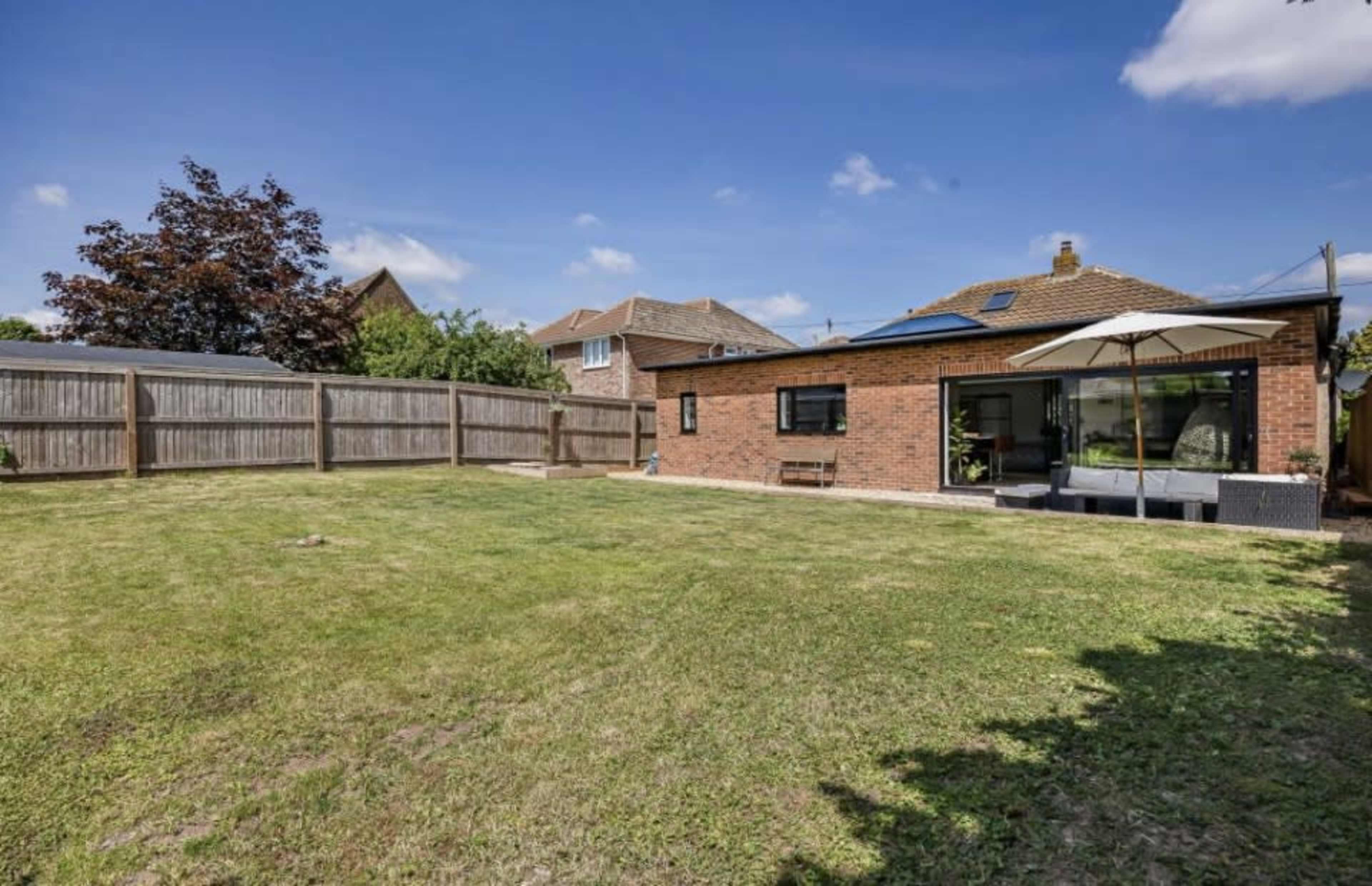 The image shows a spacious backyard with a well-maintained lawn, surrounded by a wooden fence and featuring a brick house with large windows and a patio area.