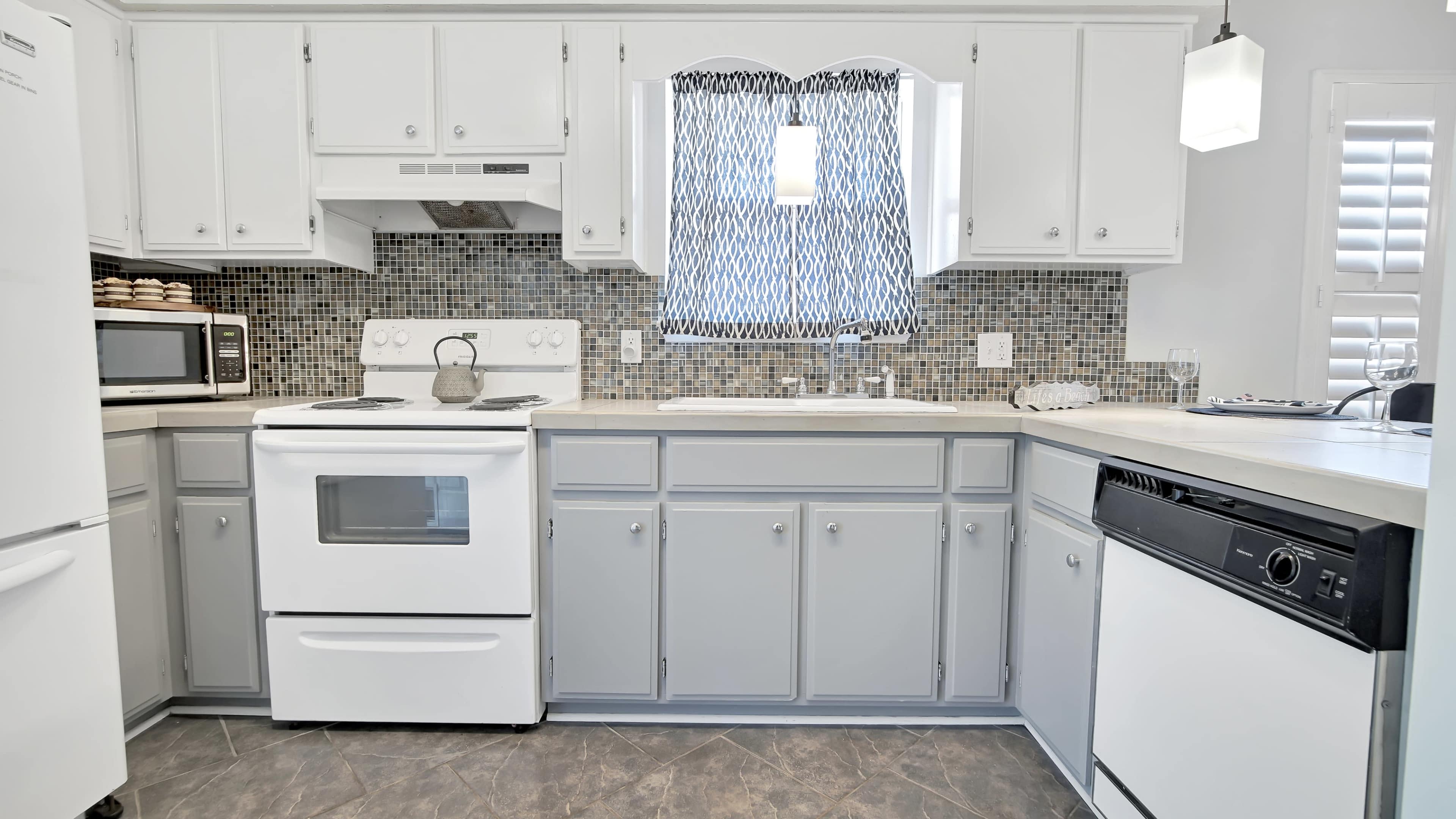 A modern kitchen with white cabinets, a gray countertop, and a tiled backsplash, featuring a stove, dishwasher, and microwave.