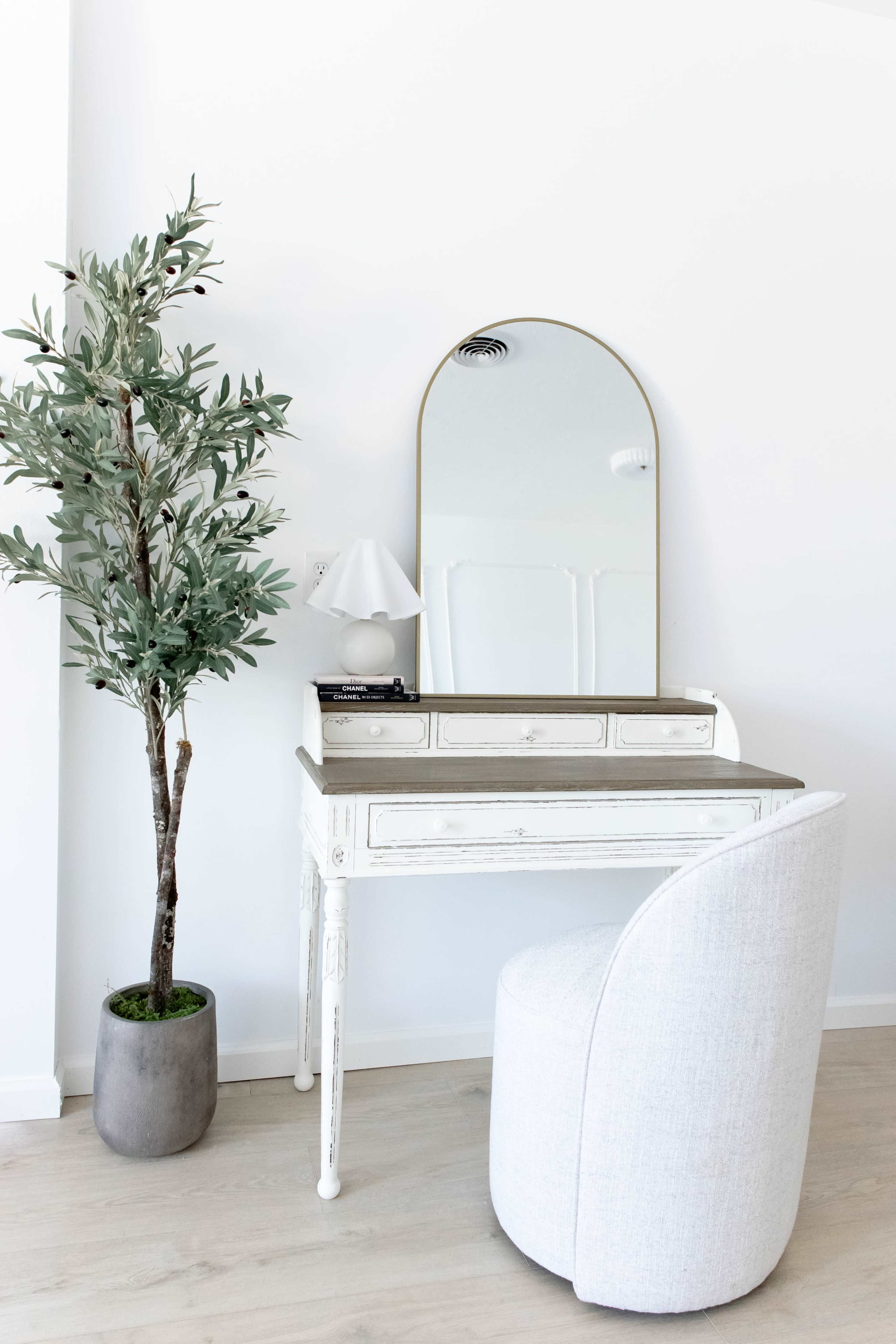 A small, elegantly designed vanity table with a round mirror is positioned next to a potted plant and a lamp in a bright room.