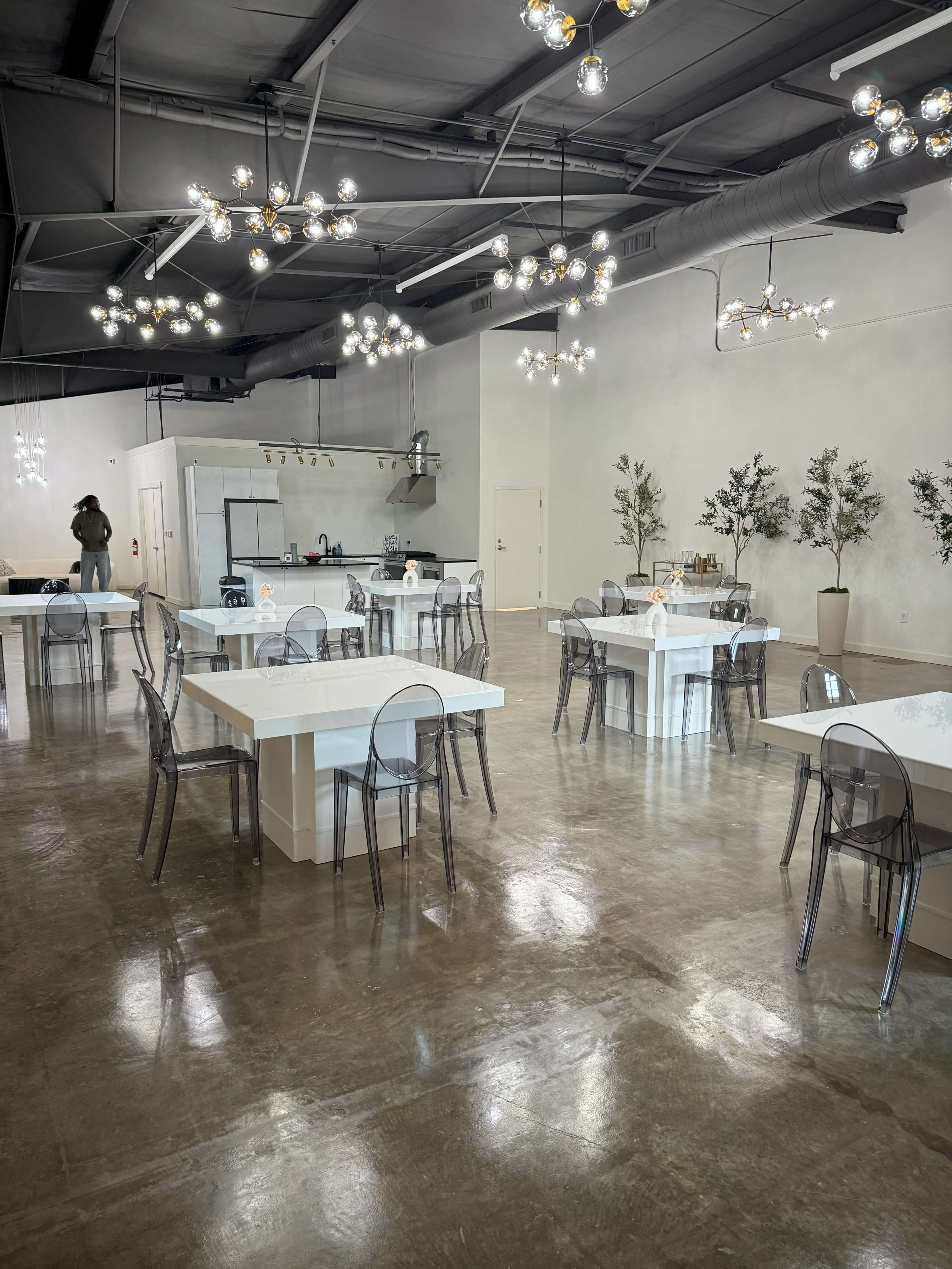 The image shows a modern, spacious dining area with multiple tables, transparent chairs, and decorative lighting fixtures.