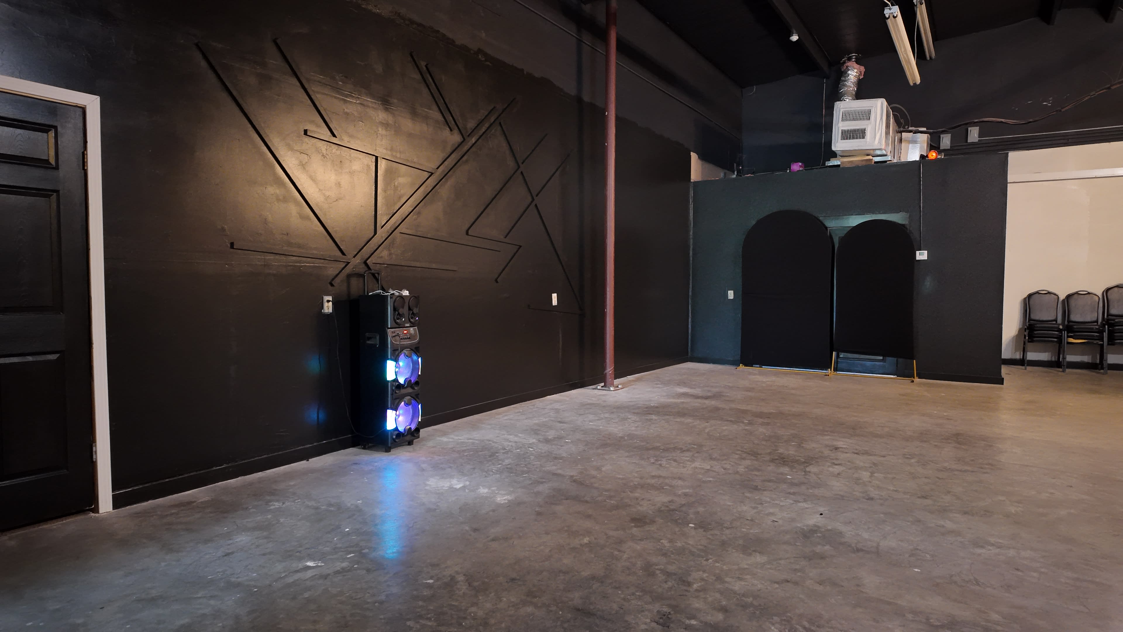 The image shows an empty room with a dark wall featuring geometric patterns, a speaker column with colored lights, and rows of chairs against a contrasting wall.