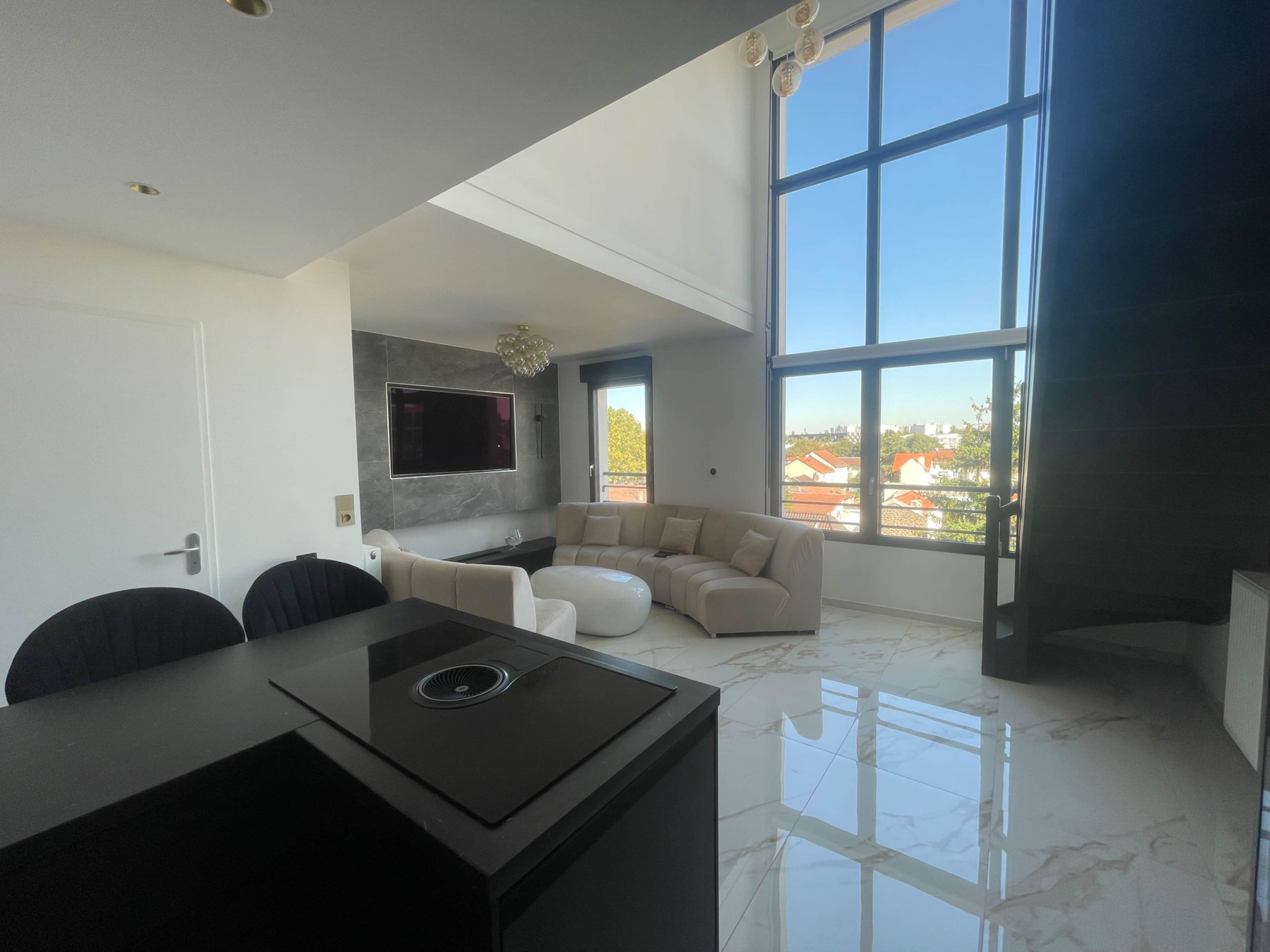 The image shows a modern living room with large windows, a light-colored sectional sofa, and a television mounted on the wall, overlooking a view of houses outside.