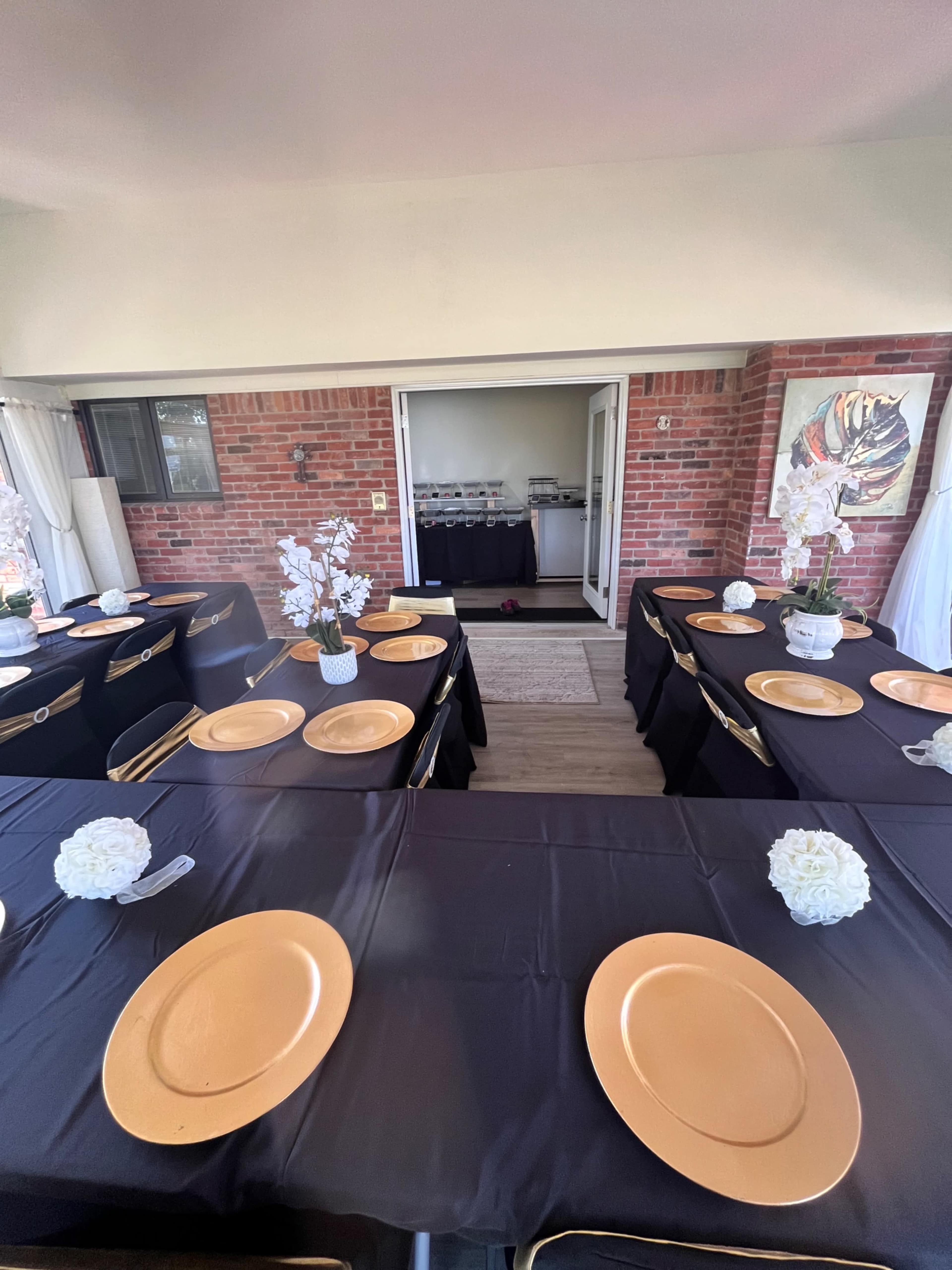 The image shows a dining area set up with black tablecloths, gold chargers, and floral centerpieces, leading to a doorway that opens into another room.