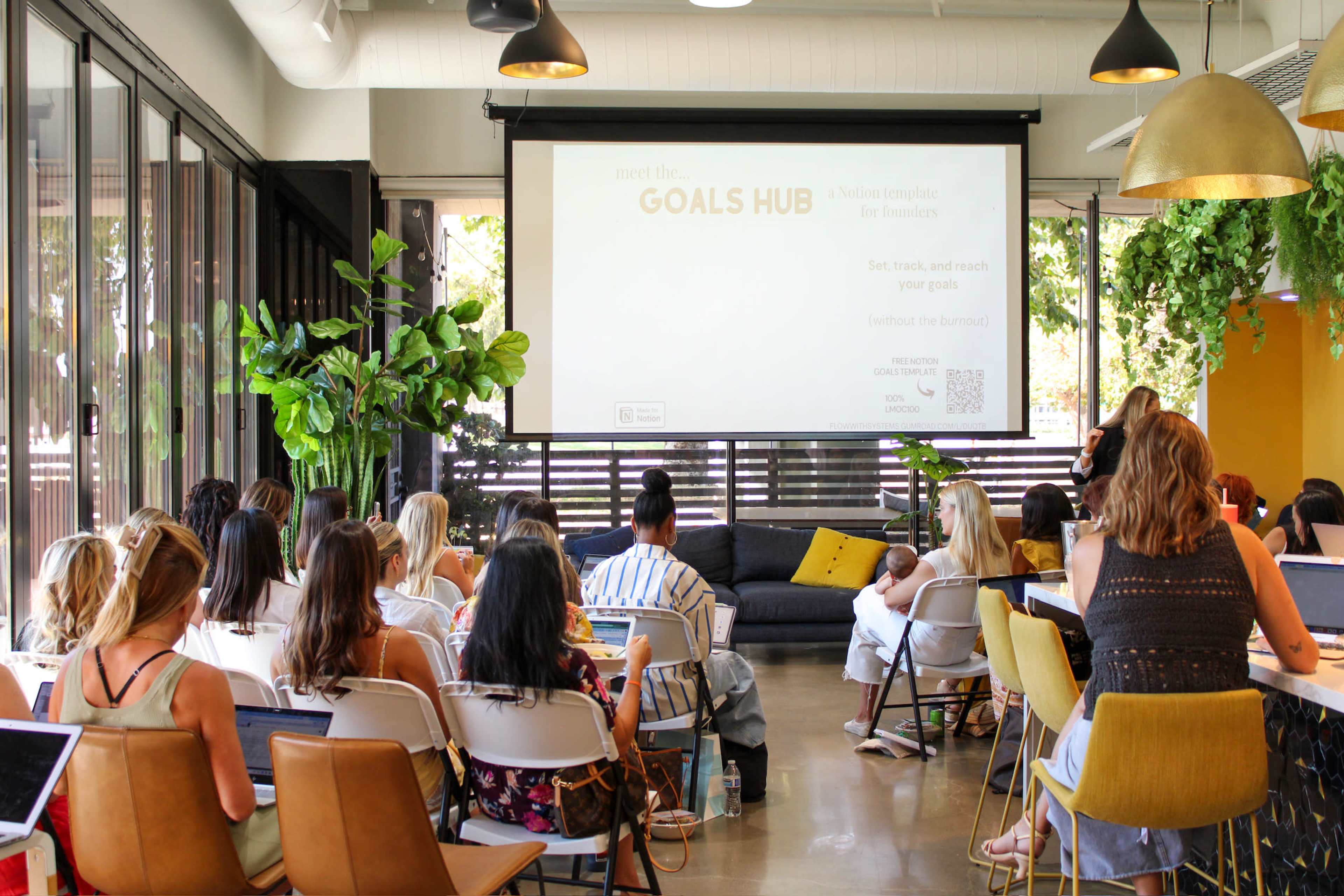 A group of people is seated in a bright, modern space with a large screen displaying a presentation about setting and achieving goals.