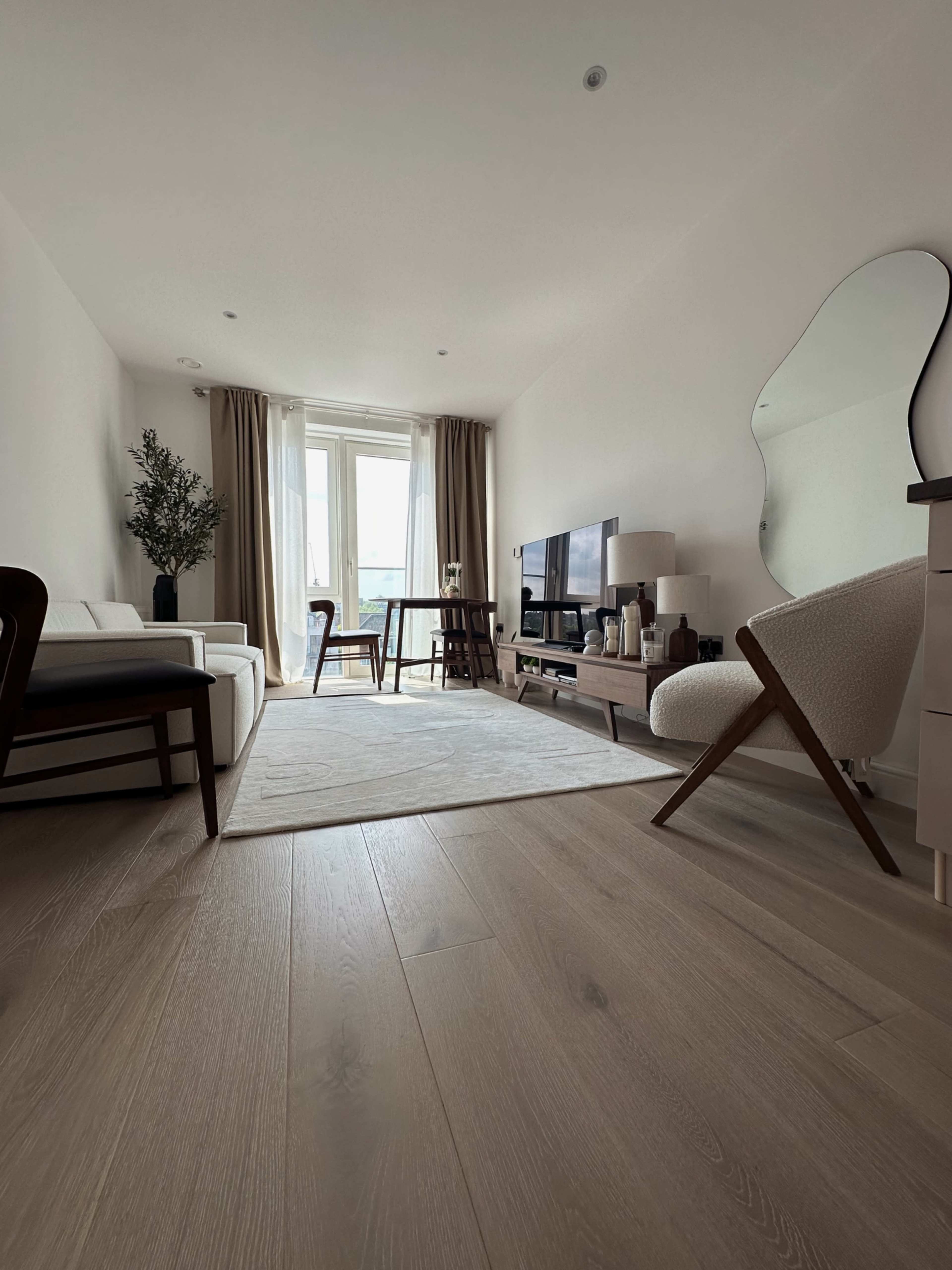 The image shows a modern living room featuring a sofa, armchair, coffee table, and television, with large windows covered by light curtains.