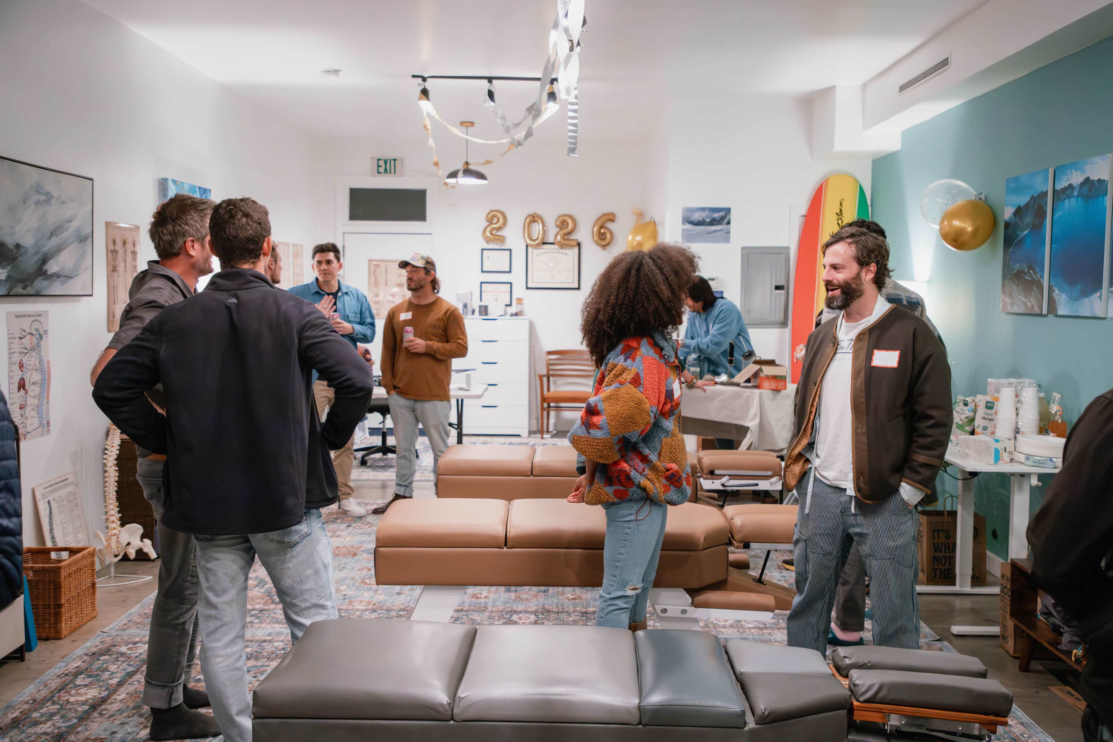 A group of people is gathered in a spacious, well-lit wellness studio with massage tables and a surfboard on display.
