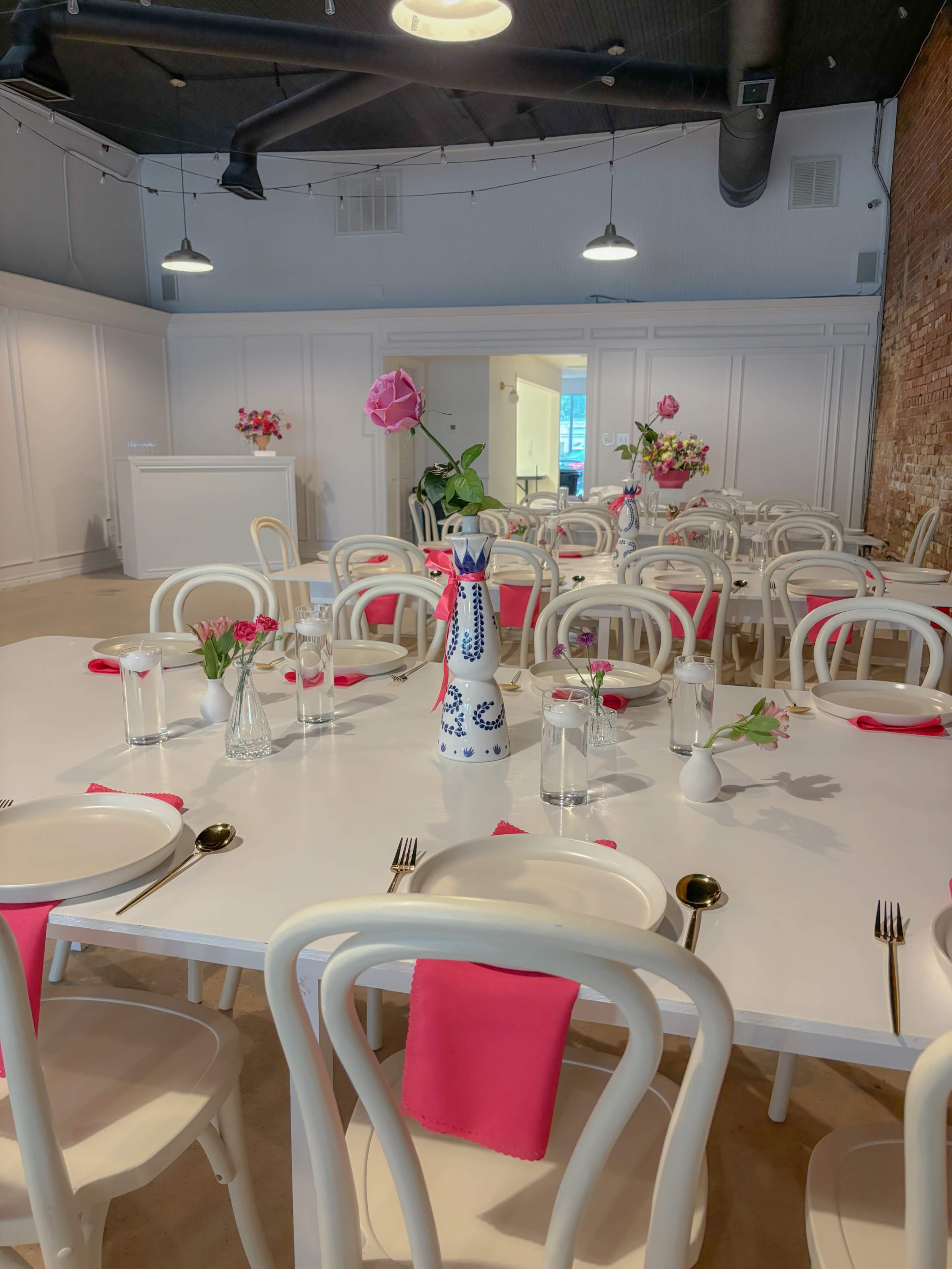 A spacious restaurant interior features white tables and chairs, with pink napkins and flower arrangements on each table.