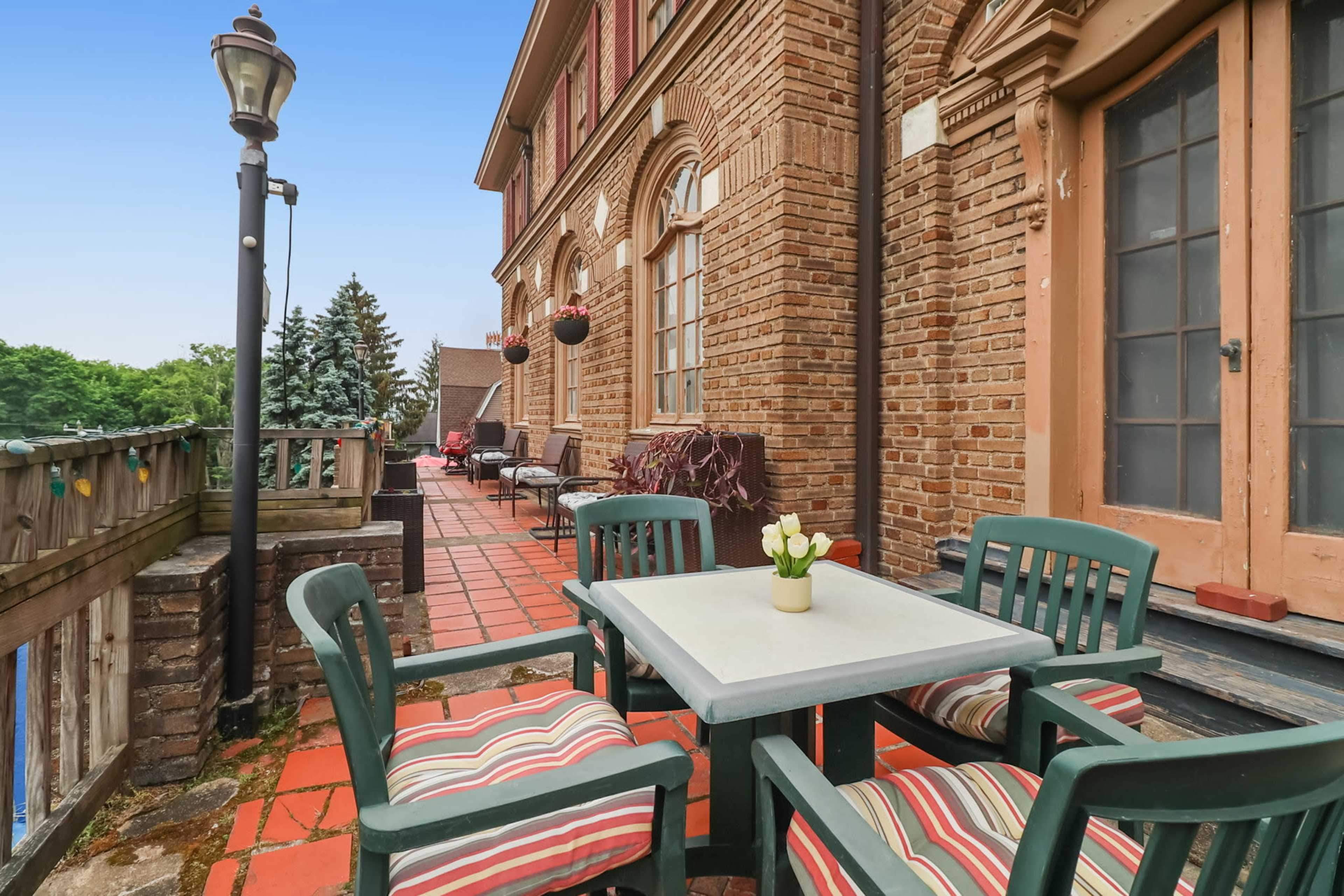 A brick building features a patio with a table and four chairs, surrounded by potted plants and a railing overlooking greenery.