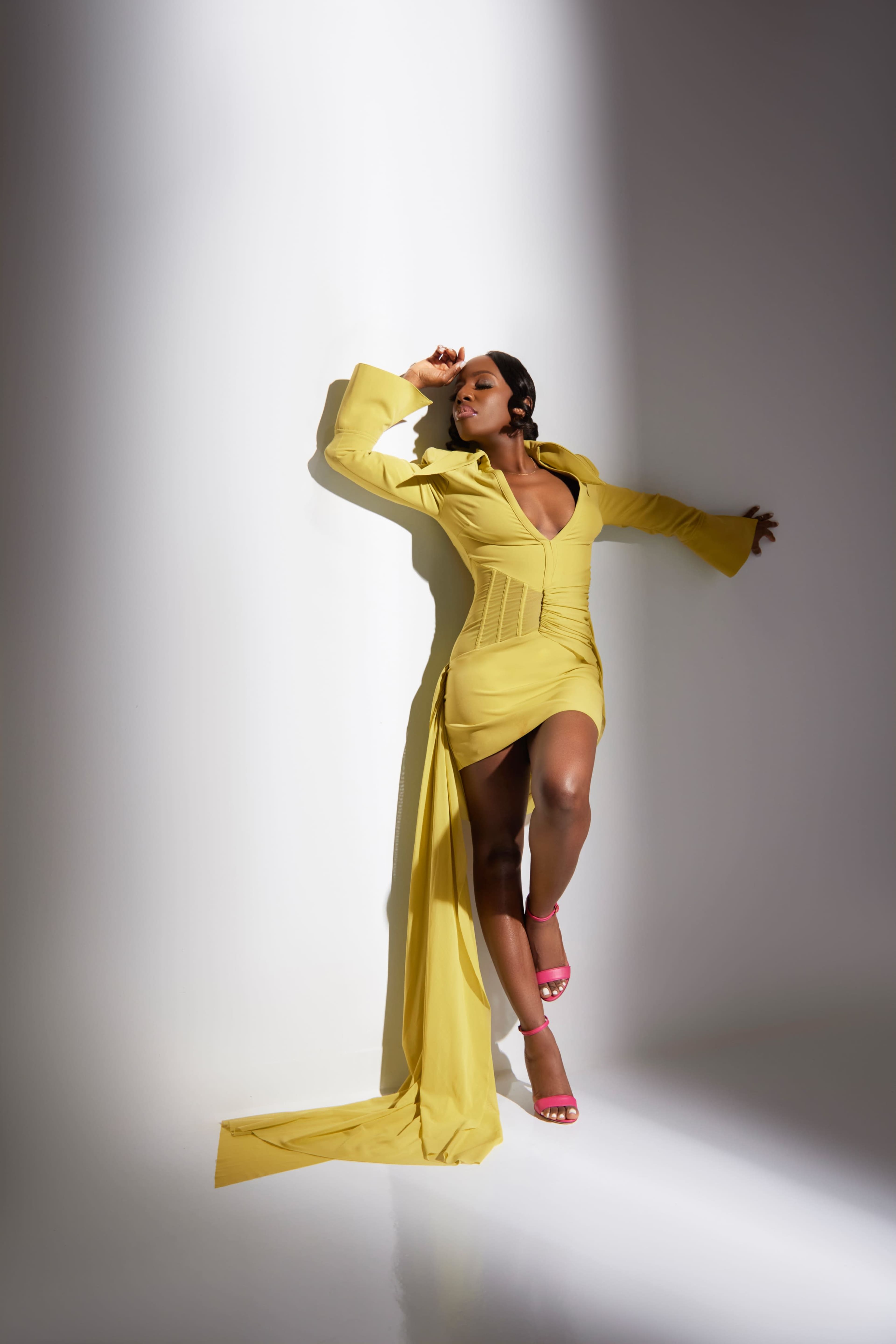 A woman poses against a simple background, wearing a yellow dress with a deep neckline and long sleeves, complemented by pink heels.