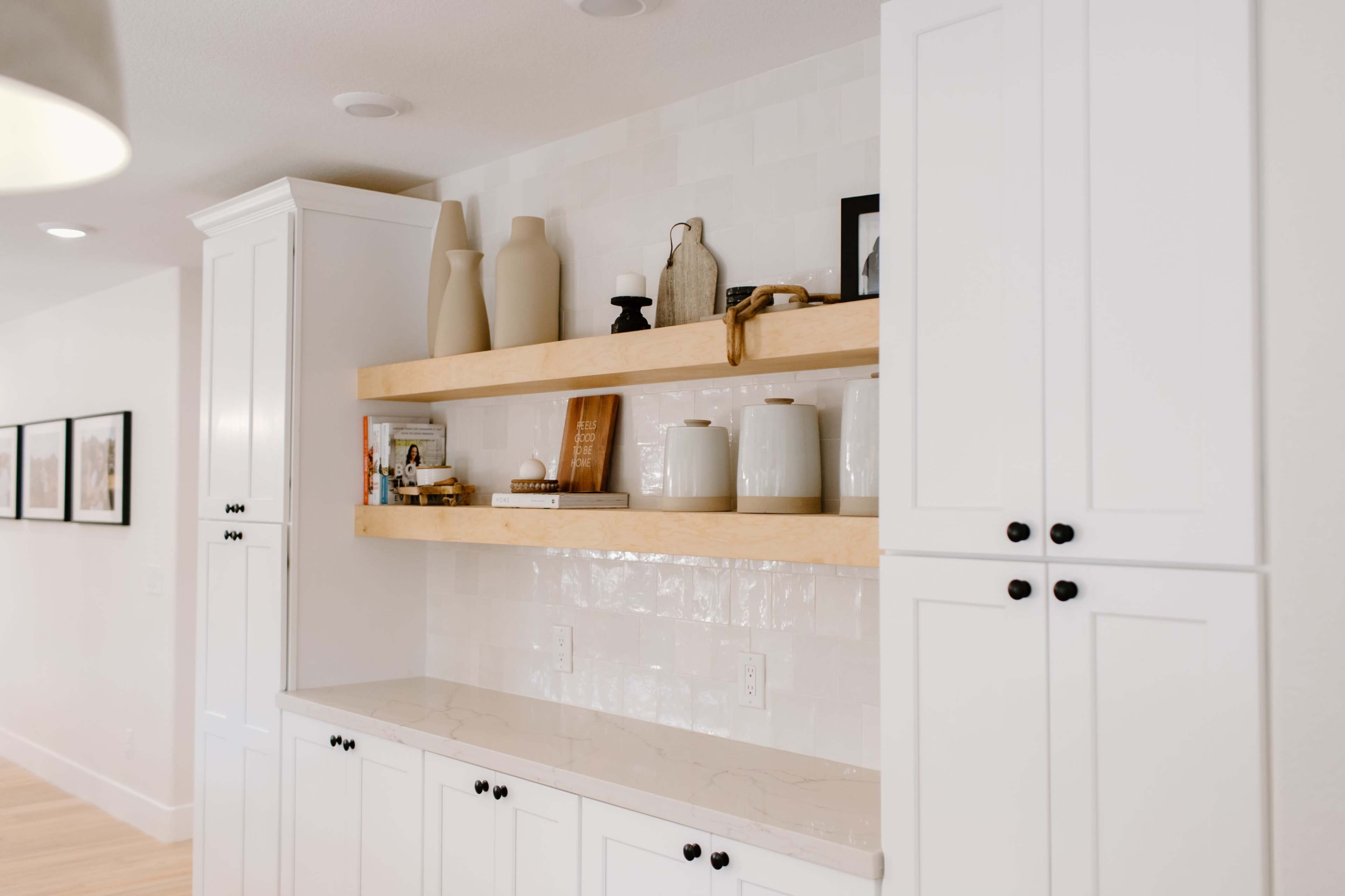 A modern kitchen space with white cabinetry, open wooden shelves displaying various decorative items, and a smooth countertop.