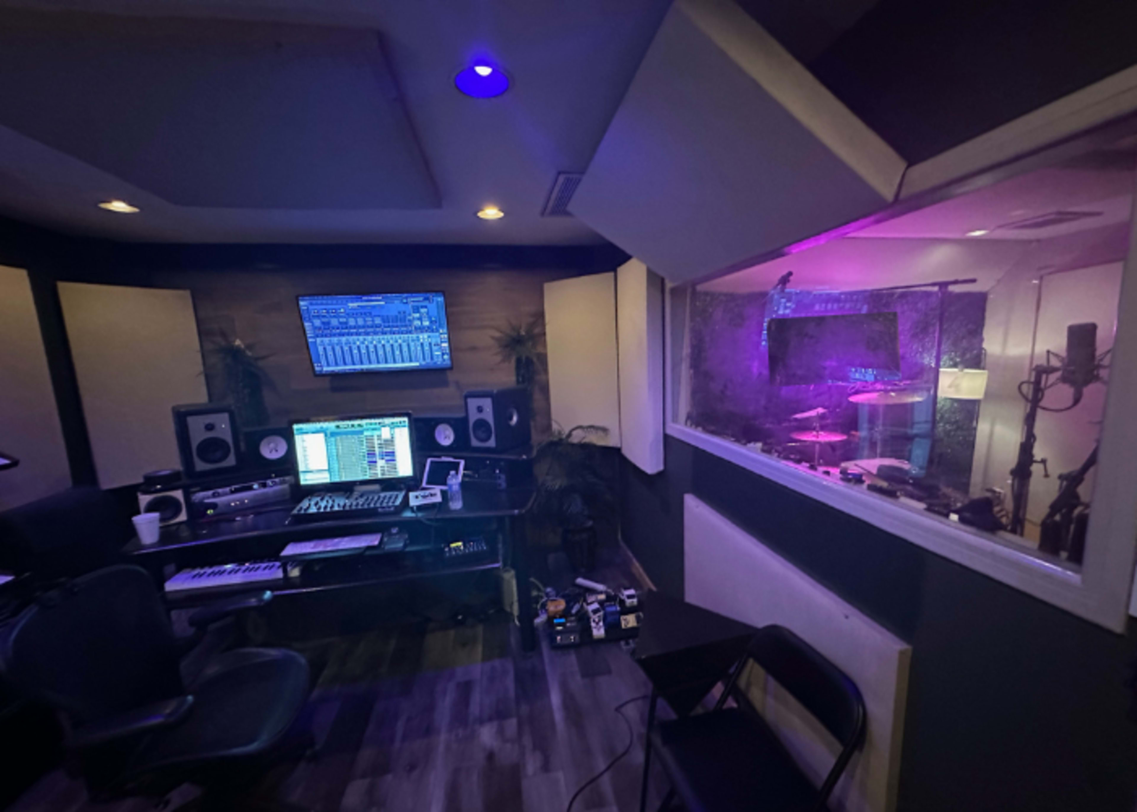 A recording studio features a control desk with monitors and equipment, adjacent to a glass-enclosed sound booth containing musical instruments.