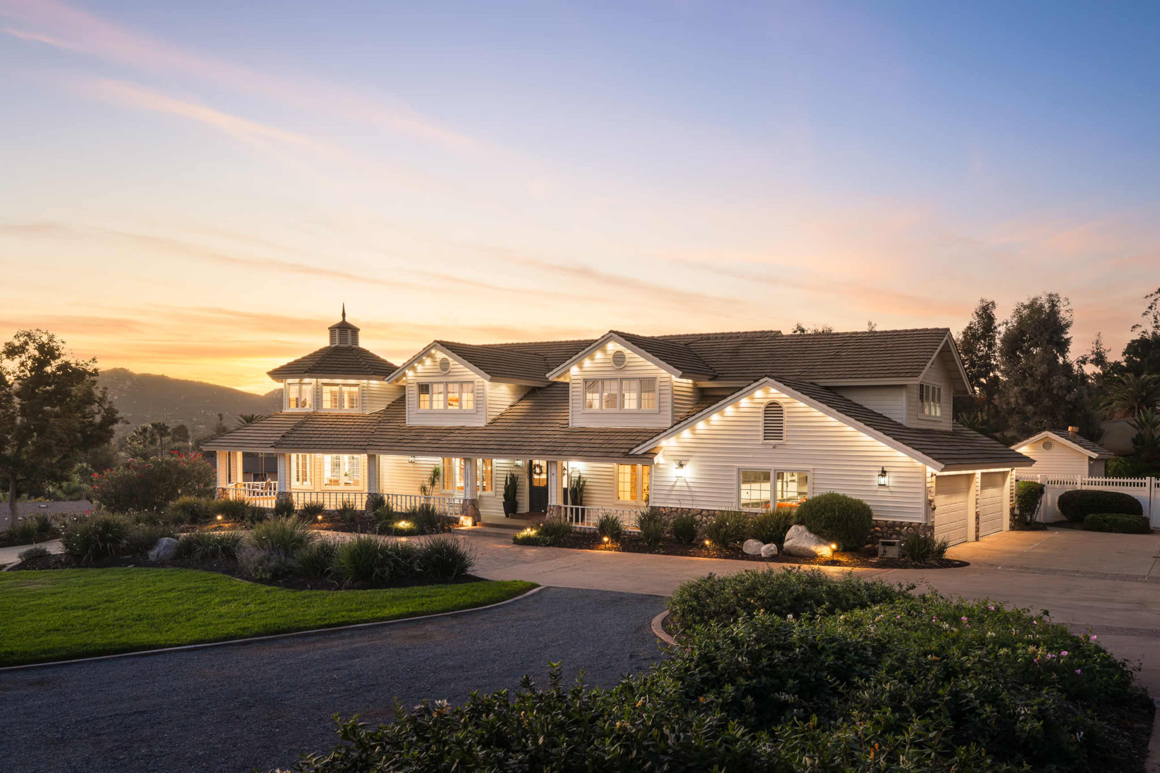 The image shows a large, modern two-story house with a white exterior, surrounded by landscaped gardens and a driveway, under a sunset sky.